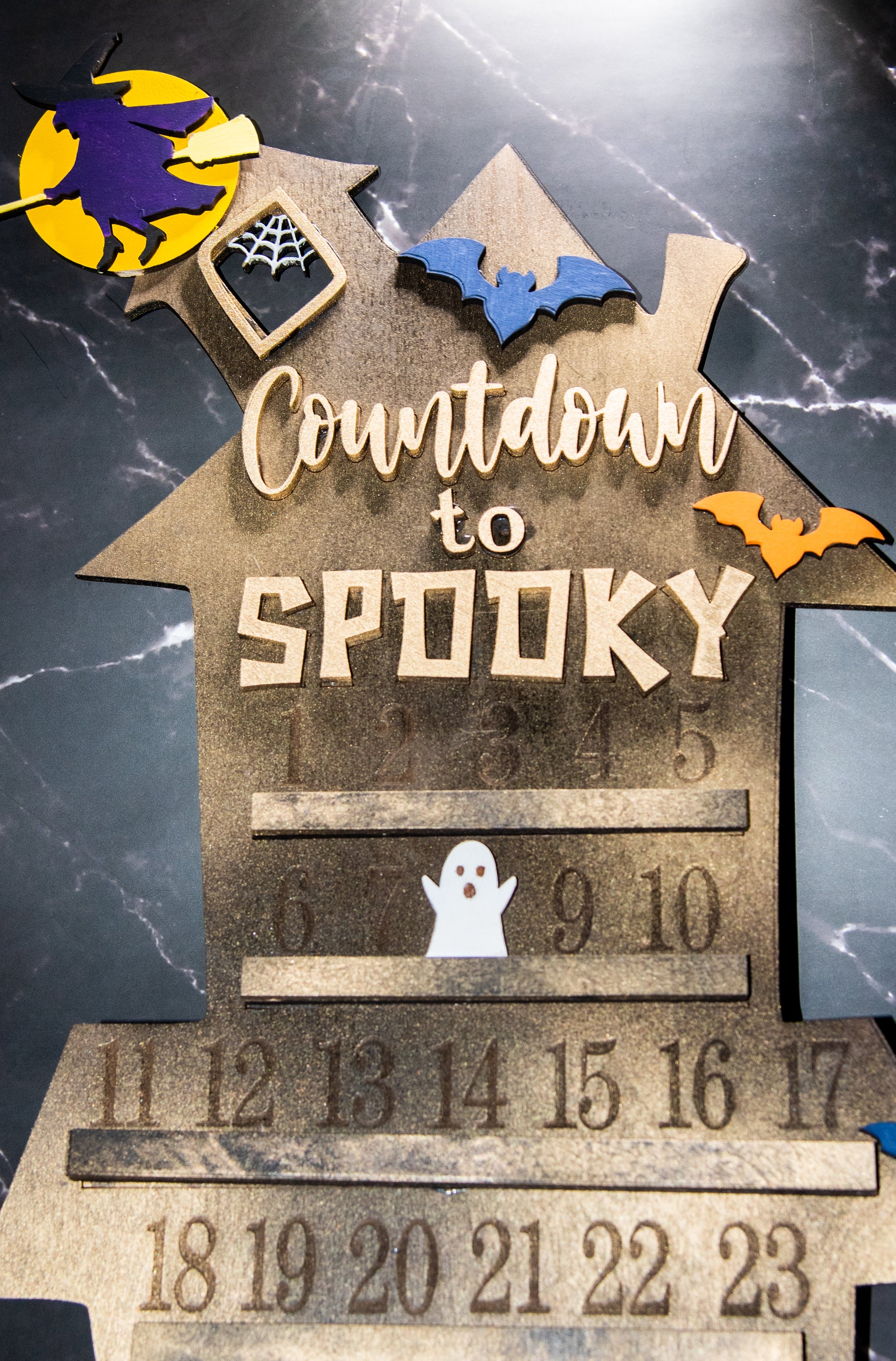 Wooden haunted house-themed Halloween countdown sign with engraved numbers from 1 to 31, painted in classic spooky colors like black, orange, and purple. Features a large haunted mansion design with bats, ghosts, and interchangeable marker icons to track the days until Halloween. Freestanding and available in two sizes, this layered wood sign is perfect for holiday decorating on a mantel, entryway, or shelf display.
