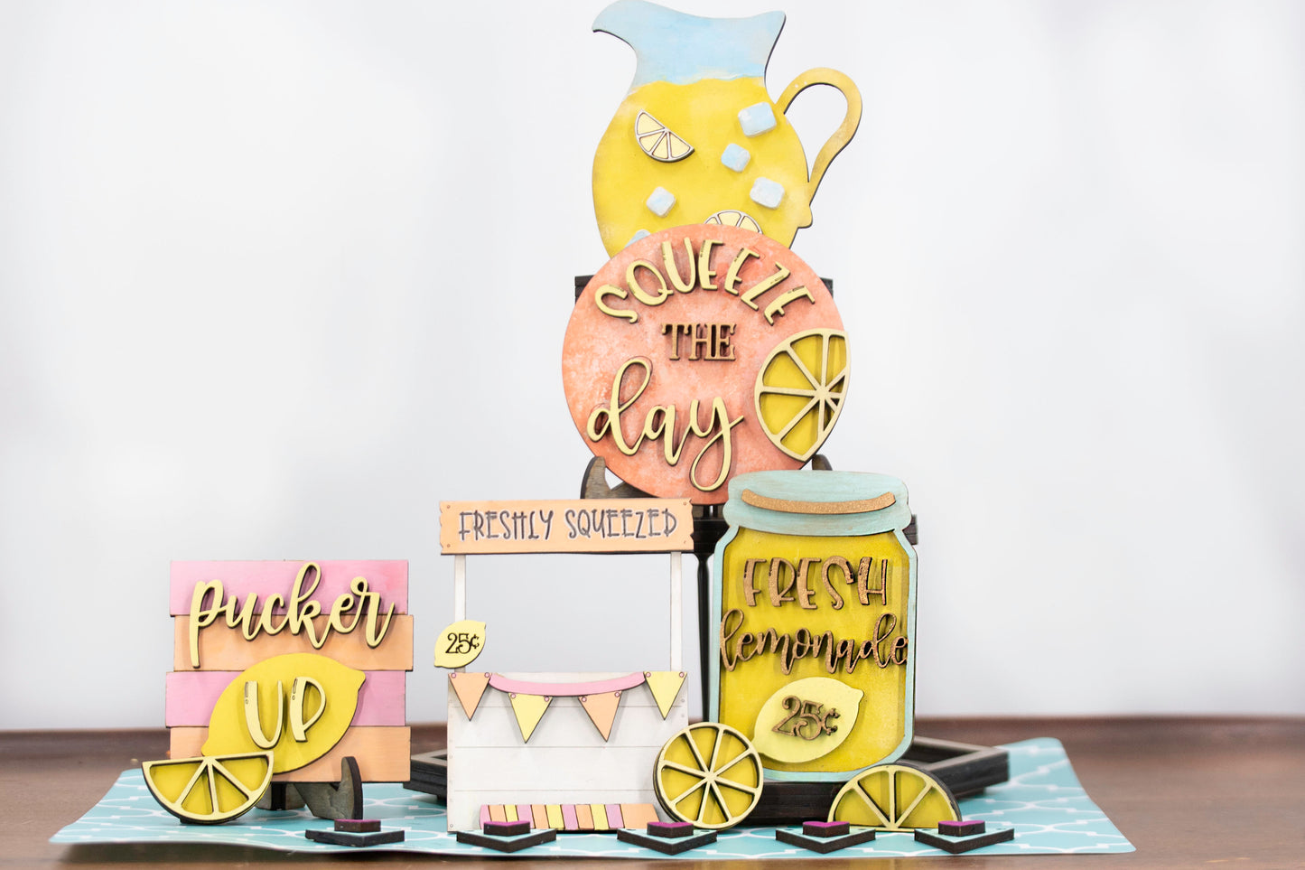 Hand-painted lemonade-themed tiered tray set featuring five wooden decor pieces in pastel pink, yellow, and orange. Includes lemon slice accents, banner flags, and stands for each item. Optional matching wooden tray base available