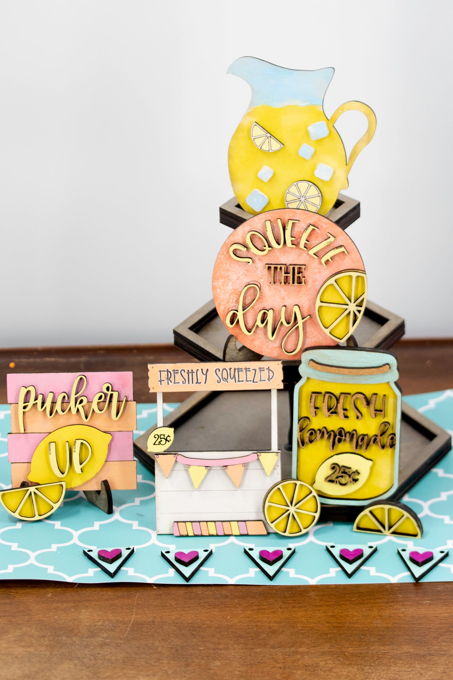 Hand-painted lemonade-themed tiered tray set featuring five wooden decor pieces in pastel pink, yellow, and orange. Includes lemon slice accents, banner flags, and stands for each item. Optional matching wooden tray base available