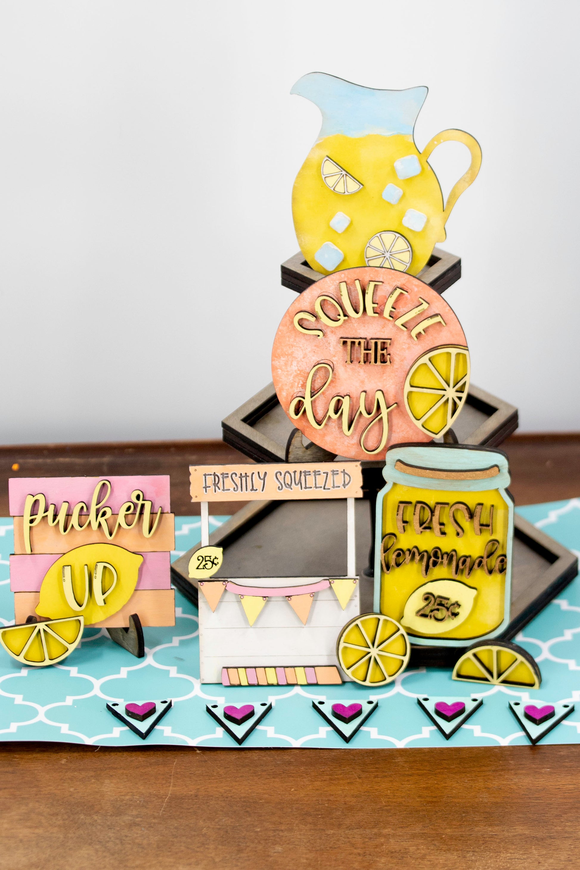 Hand-painted lemonade-themed tiered tray set featuring five wooden decor pieces in pastel pink, yellow, and orange. Includes lemon slice accents, banner flags, and stands for each item. Optional matching wooden tray base available