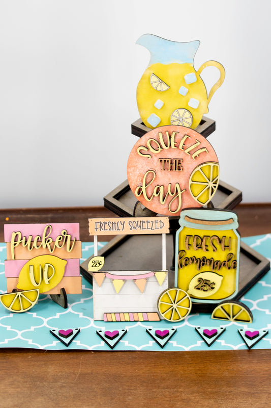 Hand-painted lemonade-themed tiered tray set featuring five wooden decor pieces in pastel pink, yellow, and orange. Includes lemon slice accents, banner flags, and stands for each item. Optional matching wooden tray base available