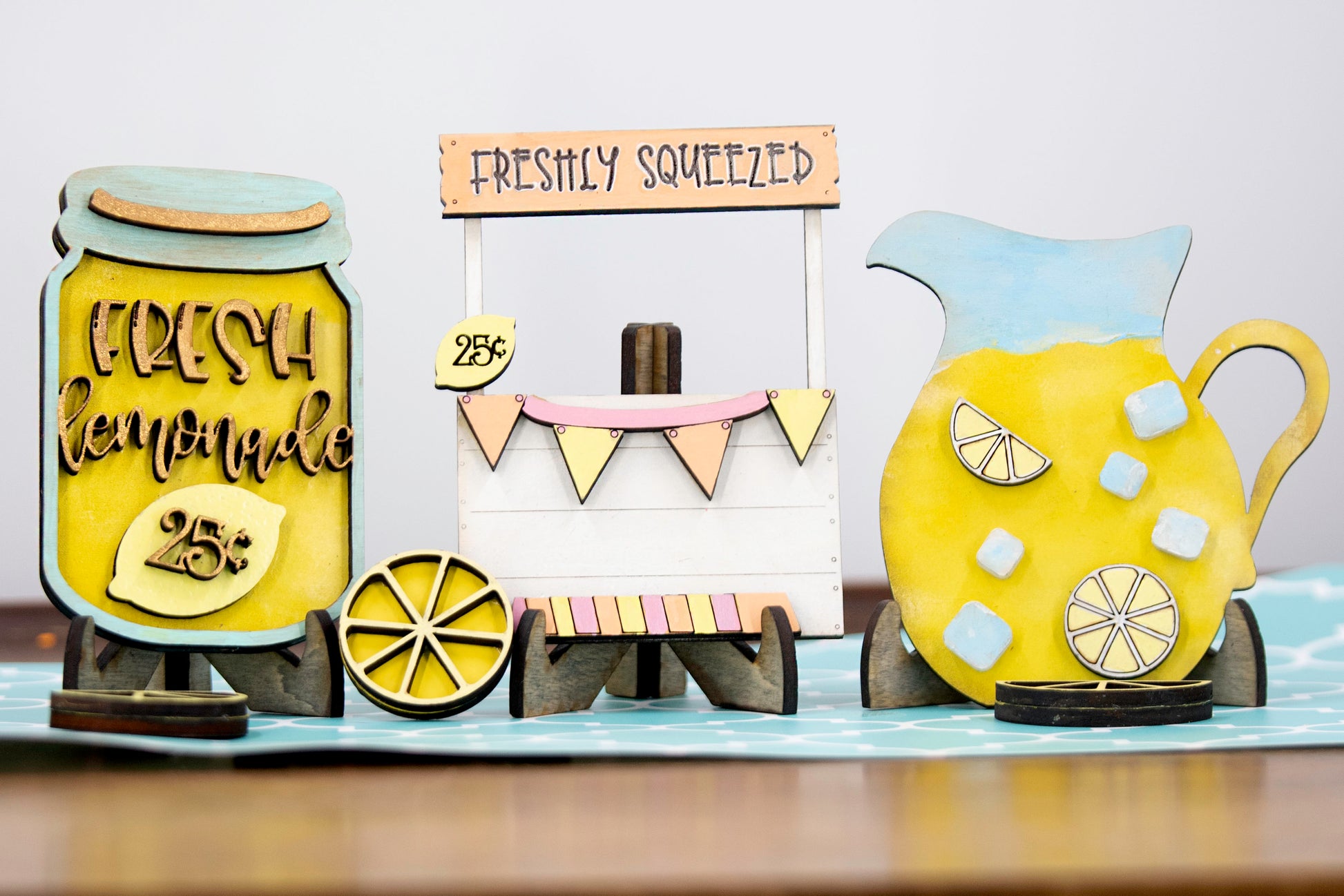 Hand-painted lemonade-themed tiered tray set featuring five wooden decor pieces in pastel pink, yellow, and orange. Includes lemon slice accents, banner flags, and stands for each item. Optional matching wooden tray base available