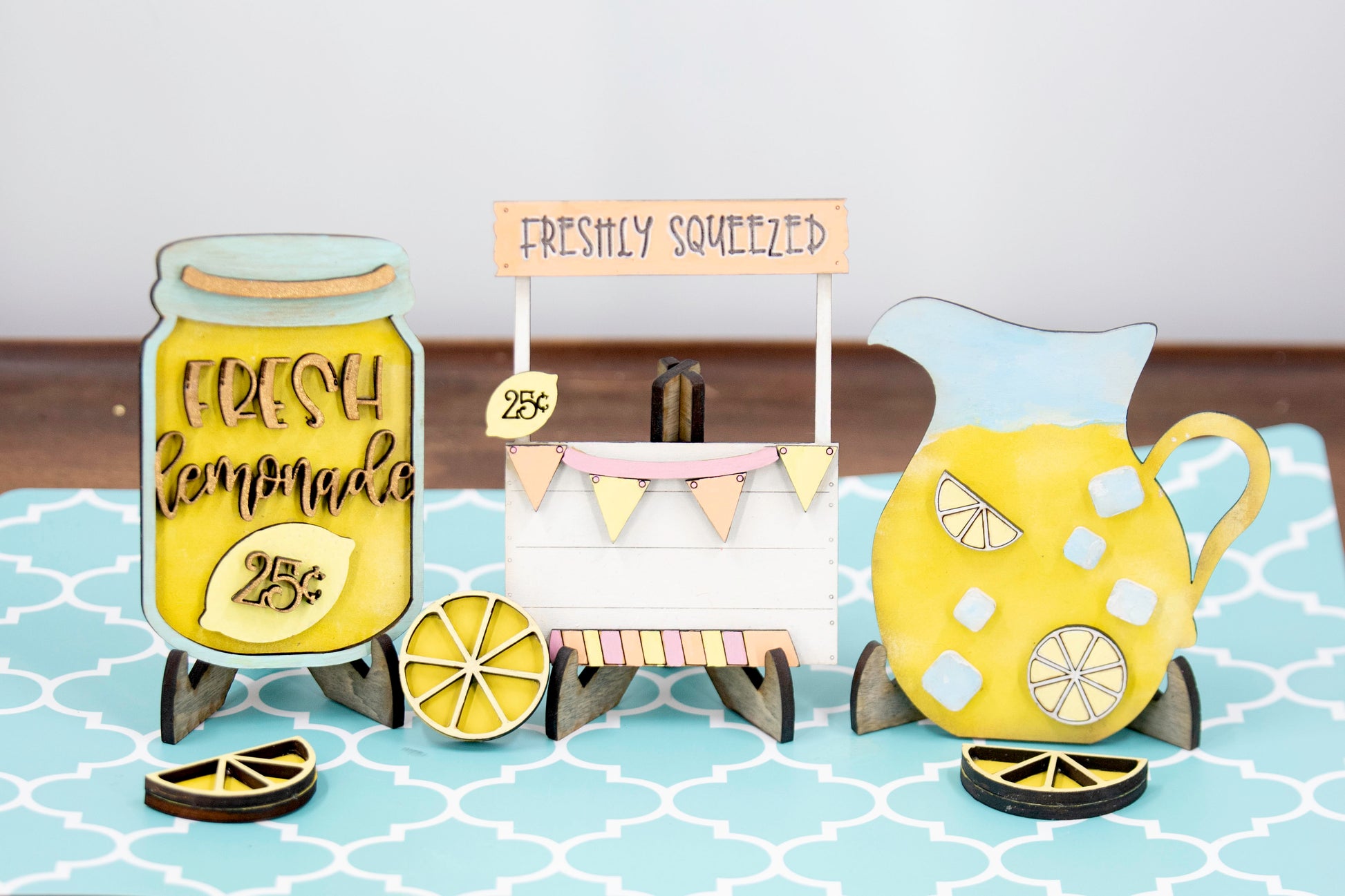 Hand-painted lemonade-themed tiered tray set featuring five wooden decor pieces in pastel pink, yellow, and orange. Includes lemon slice accents, banner flags, and stands for each item. Optional matching wooden tray base available