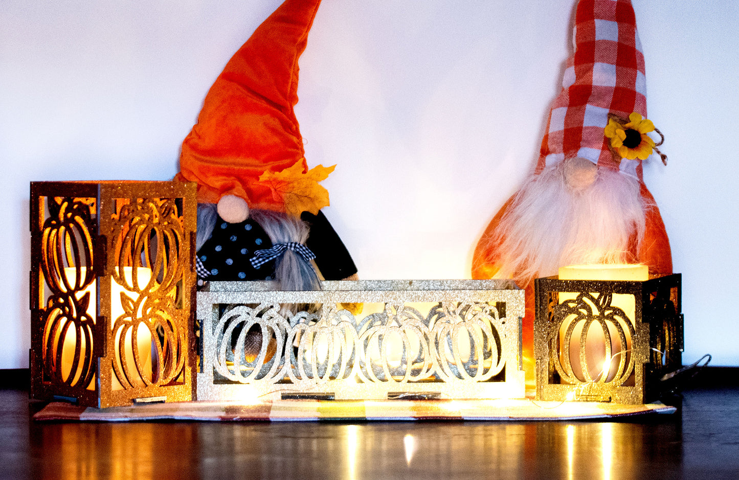 et of three intricately designed metal and wood pumpkin candle holders displayed on a checkered fall table runner. The set includes a tall rectangular lantern, a horizontal centerpiece tray, and a small square holder, each featuring layered pumpkin cutouts that beautifully glow with the warm light of tea candles inside—perfect for cozy fall, Thanksgiving, or farmhouse-style decor.