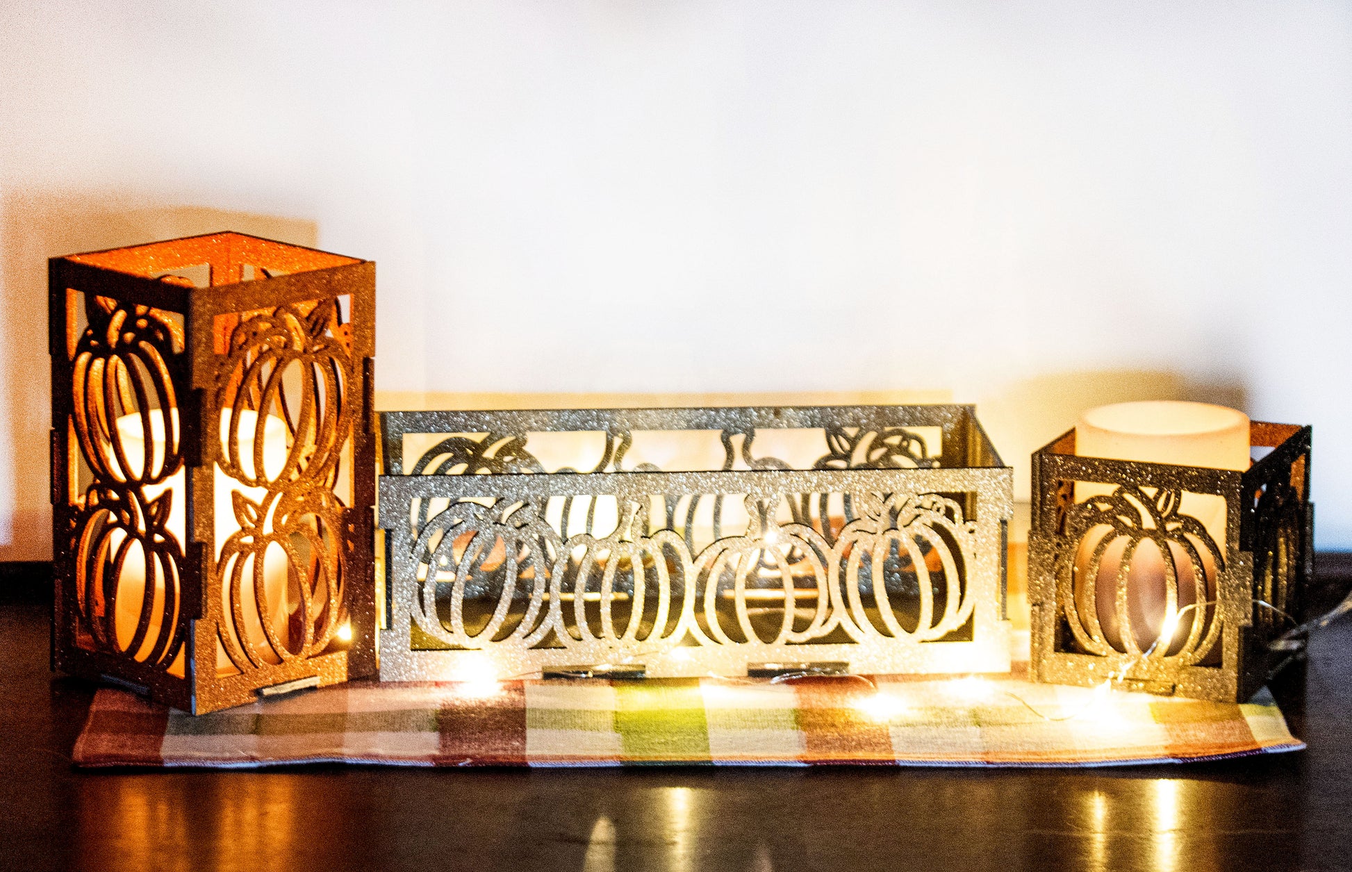et of three intricately designed metal and wood pumpkin candle holders displayed on a checkered fall table runner. The set includes a tall rectangular lantern, a horizontal centerpiece tray, and a small square holder, each featuring layered pumpkin cutouts that beautifully glow with the warm light of tea candles inside—perfect for cozy fall, Thanksgiving, or farmhouse-style decor.