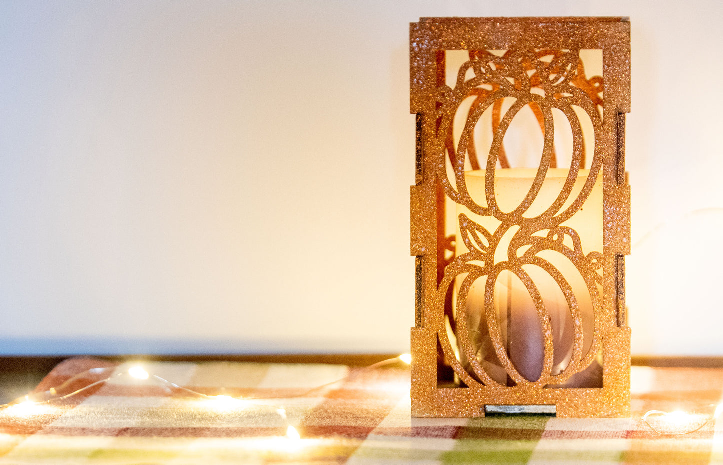 et of three intricately designed metal and wood pumpkin candle holders displayed on a checkered fall table runner. The set includes a tall rectangular lantern, a horizontal centerpiece tray, and a small square holder, each featuring layered pumpkin cutouts that beautifully glow with the warm light of tea candles inside—perfect for cozy fall, Thanksgiving, or farmhouse-style decor.