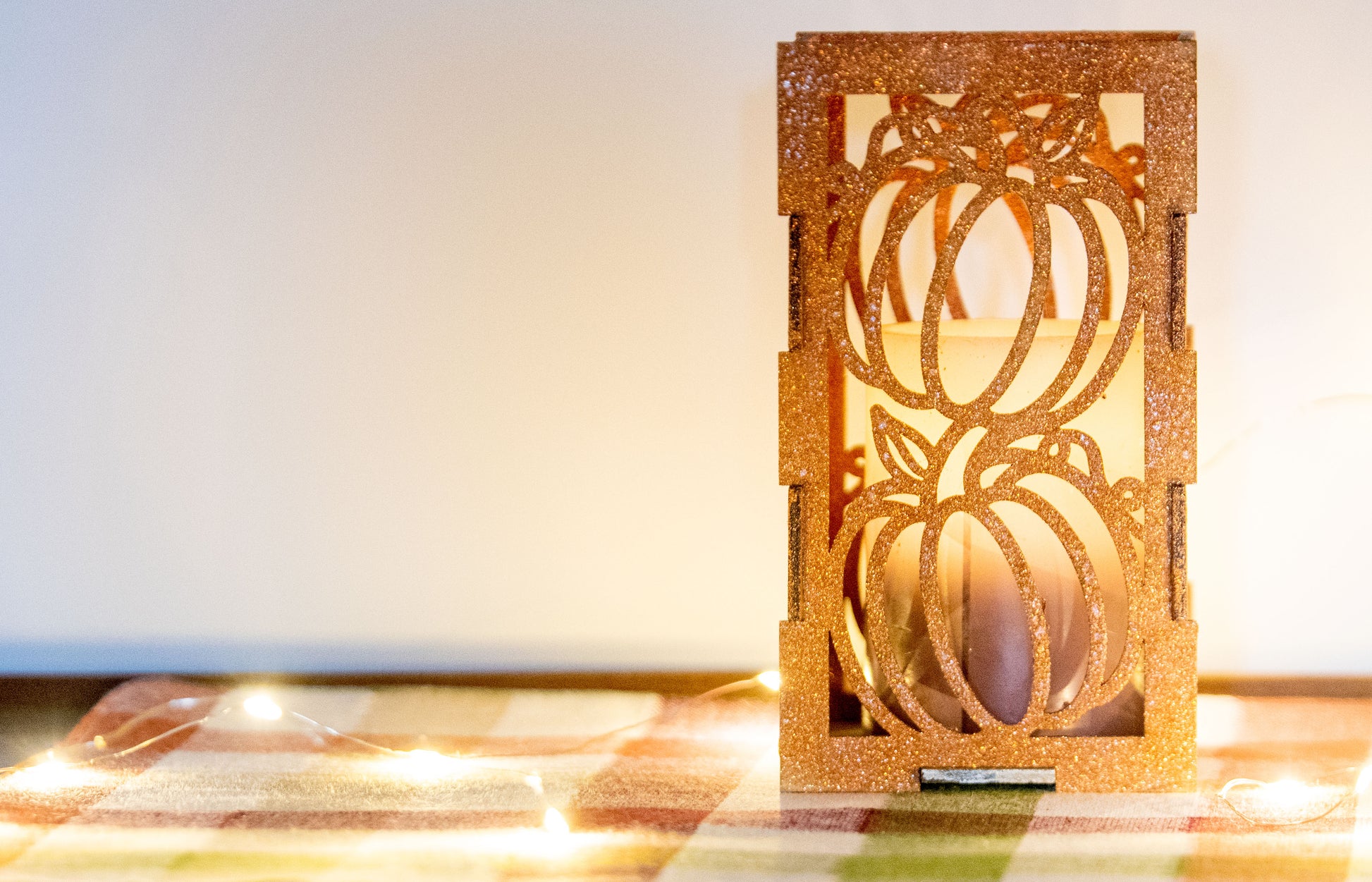 et of three intricately designed metal and wood pumpkin candle holders displayed on a checkered fall table runner. The set includes a tall rectangular lantern, a horizontal centerpiece tray, and a small square holder, each featuring layered pumpkin cutouts that beautifully glow with the warm light of tea candles inside—perfect for cozy fall, Thanksgiving, or farmhouse-style decor.