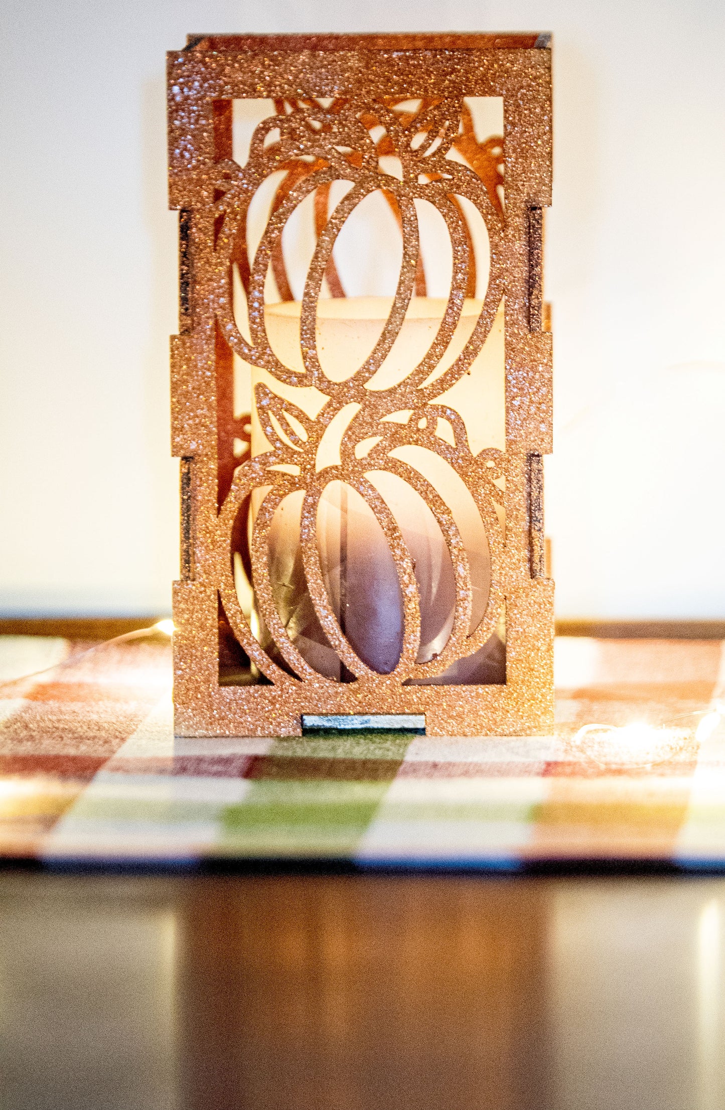 et of three intricately designed metal and wood pumpkin candle holders displayed on a checkered fall table runner. The set includes a tall rectangular lantern, a horizontal centerpiece tray, and a small square holder, each featuring layered pumpkin cutouts that beautifully glow with the warm light of tea candles inside—perfect for cozy fall, Thanksgiving, or farmhouse-style decor.