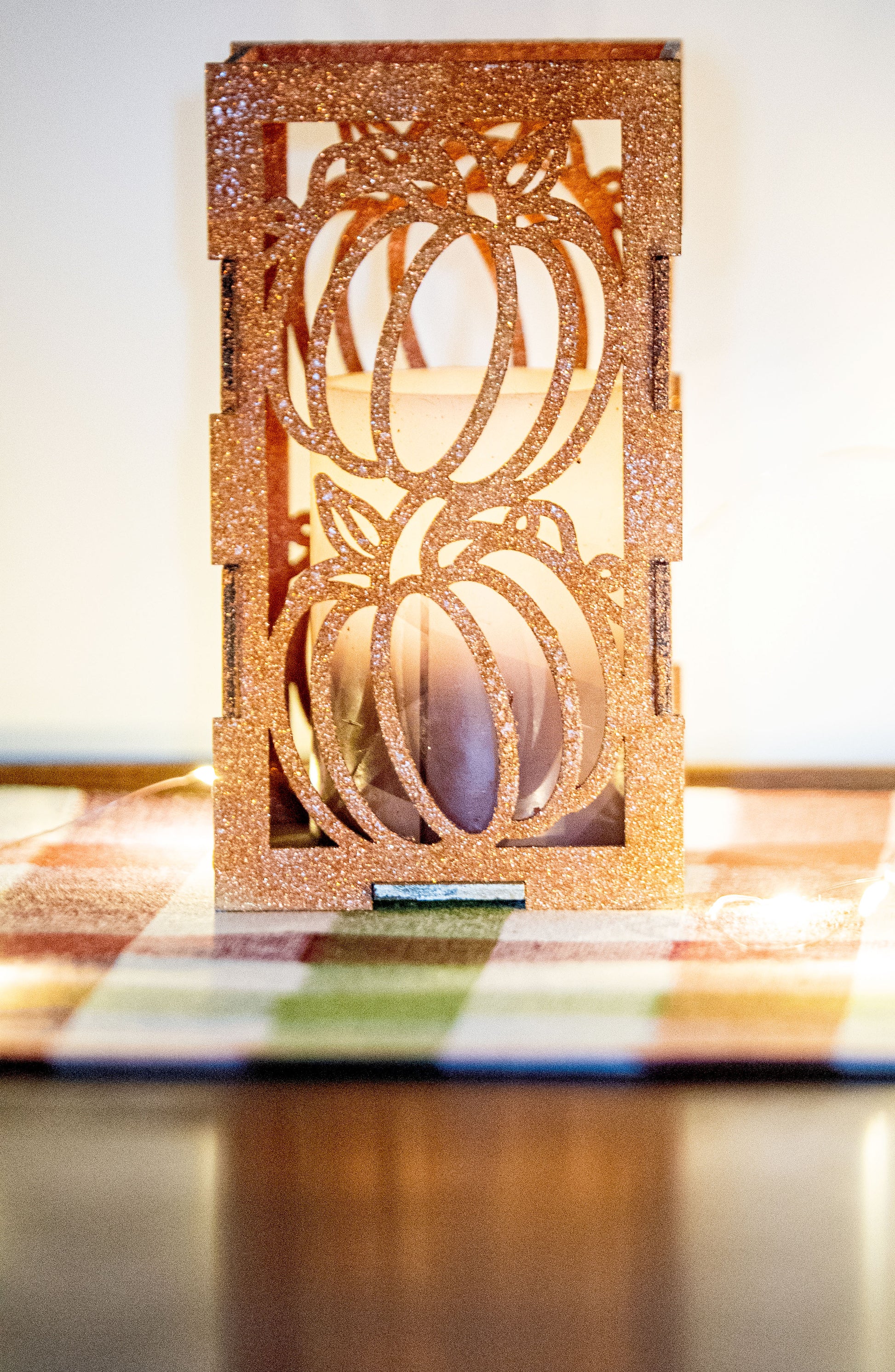 et of three intricately designed metal and wood pumpkin candle holders displayed on a checkered fall table runner. The set includes a tall rectangular lantern, a horizontal centerpiece tray, and a small square holder, each featuring layered pumpkin cutouts that beautifully glow with the warm light of tea candles inside—perfect for cozy fall, Thanksgiving, or farmhouse-style decor.