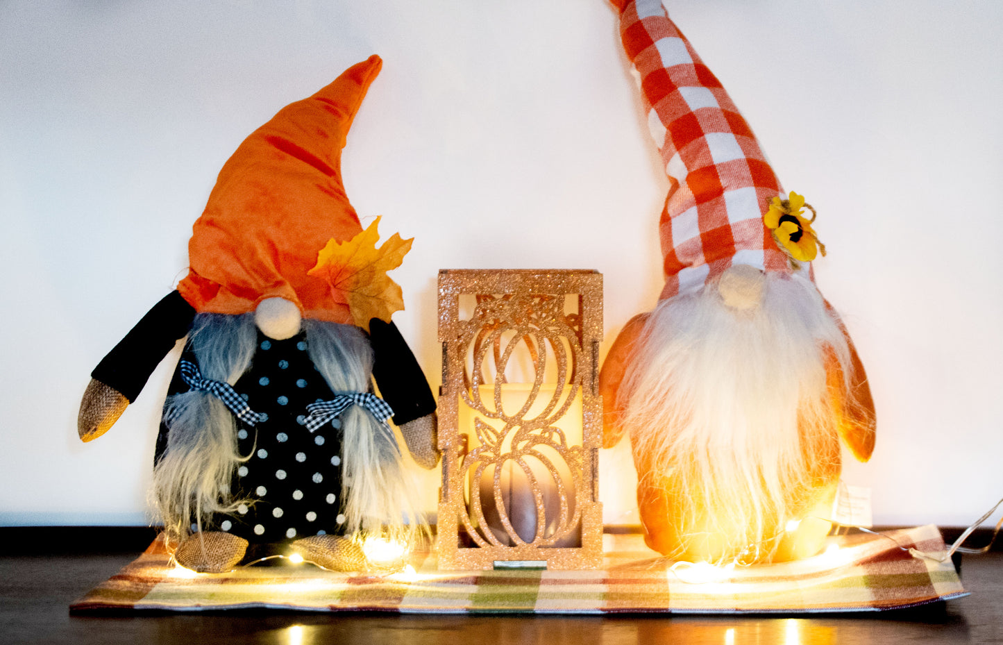 et of three intricately designed metal and wood pumpkin candle holders displayed on a checkered fall table runner. The set includes a tall rectangular lantern, a horizontal centerpiece tray, and a small square holder, each featuring layered pumpkin cutouts that beautifully glow with the warm light of tea candles inside—perfect for cozy fall, Thanksgiving, or farmhouse-style decor.