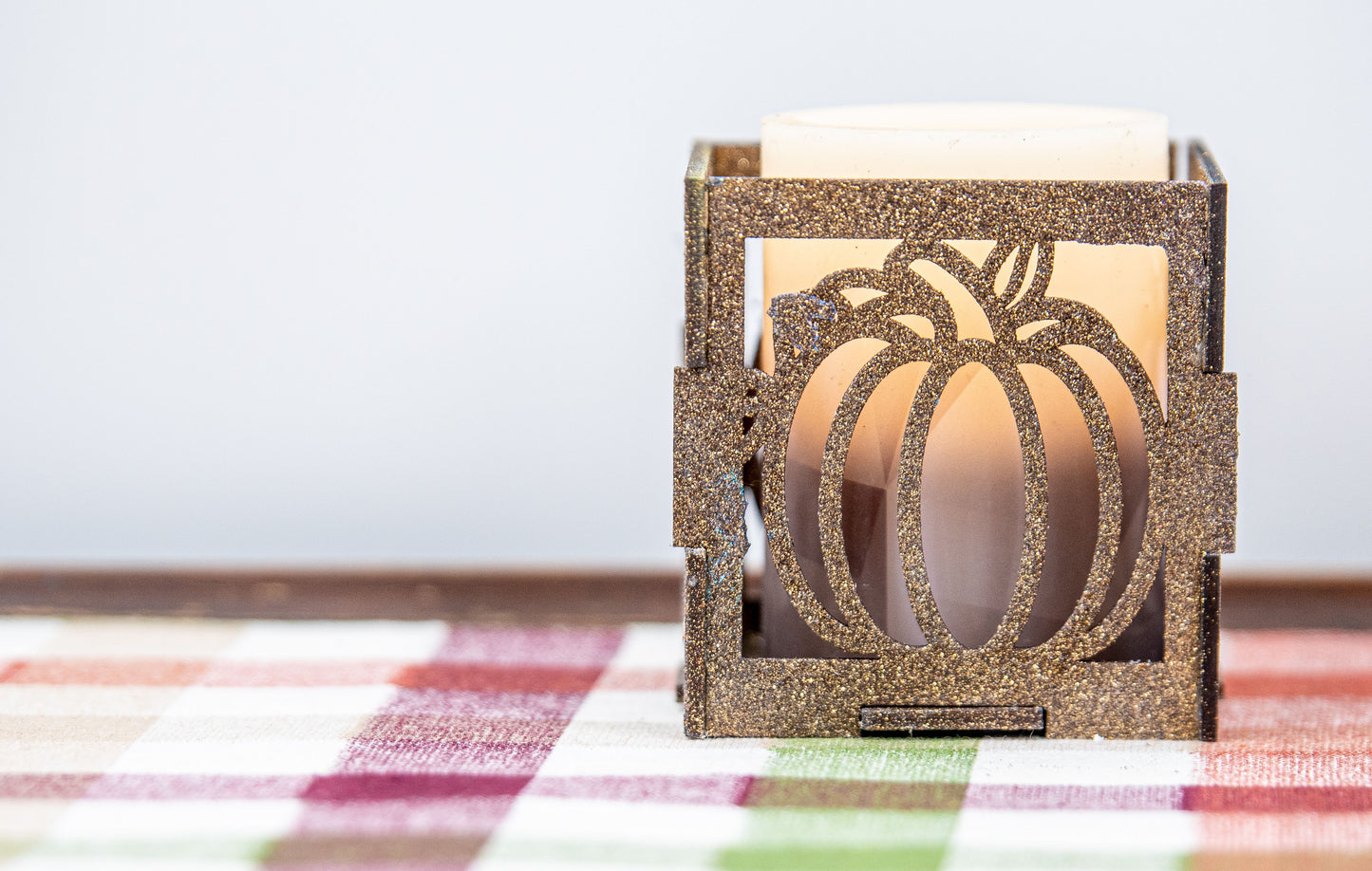 et of three intricately designed metal and wood pumpkin candle holders displayed on a checkered fall table runner. The set includes a tall rectangular lantern, a horizontal centerpiece tray, and a small square holder, each featuring layered pumpkin cutouts that beautifully glow with the warm light of tea candles inside—perfect for cozy fall, Thanksgiving, or farmhouse-style decor.
