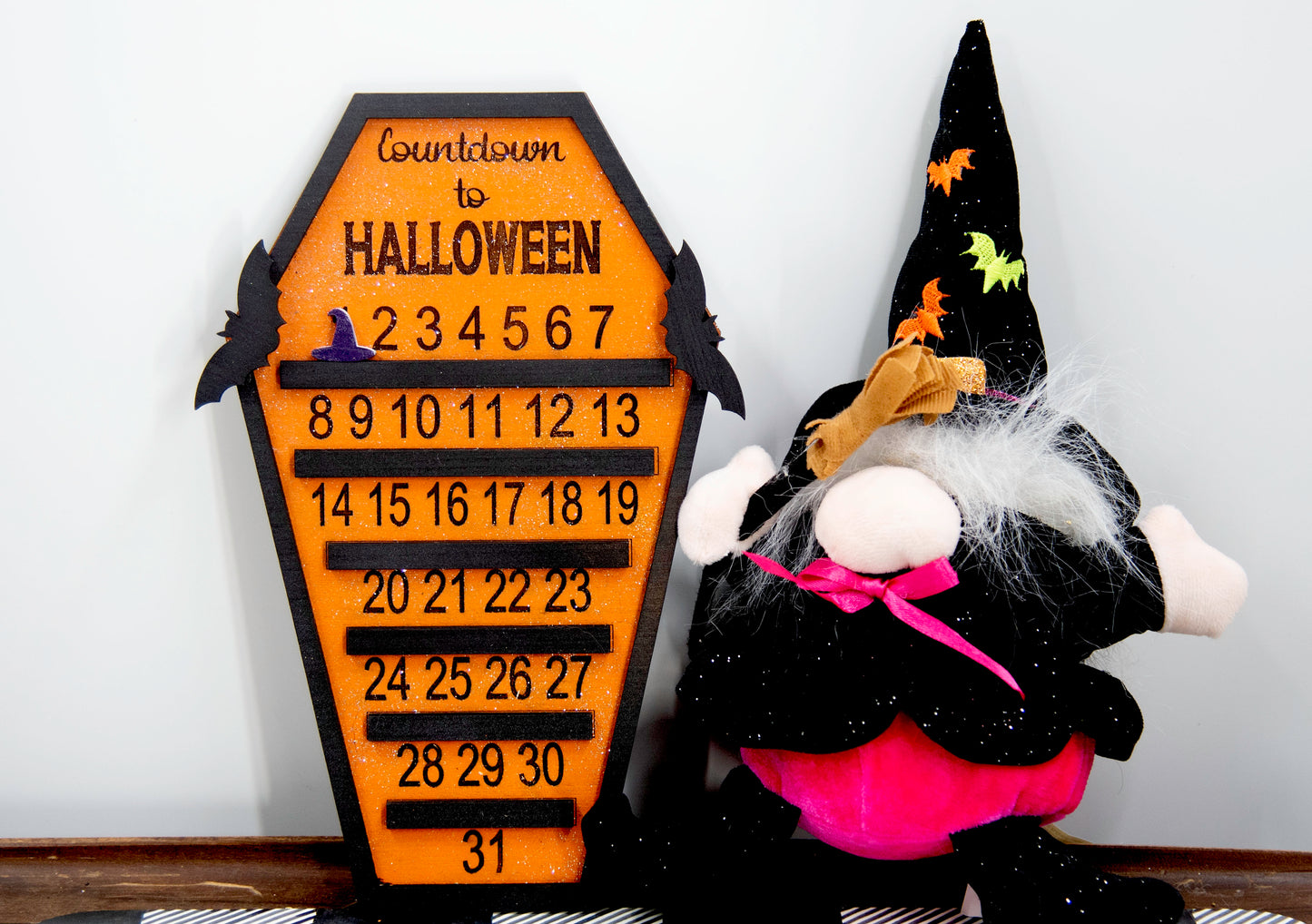 Wooden Halloween countdown sign shaped like a coffin with an orange glitter background, black trim frame, engraved 'Countdown to Halloween' at the top, engraved numbers 1–31 with small black shelves below each, and three interchangeable markers: ghost, witch hat, and pumpkin. Includes bat accents on top.