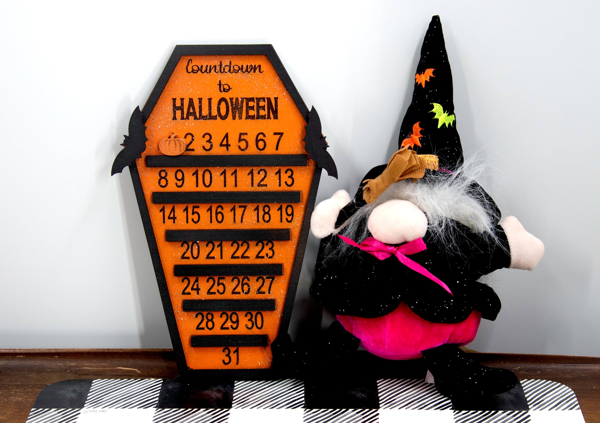 Wooden Halloween countdown sign shaped like a coffin with an orange glitter background, black trim frame, engraved 'Countdown to Halloween' at the top, engraved numbers 1–31 with small black shelves below each, and three interchangeable markers: ghost, witch hat, and pumpkin. Includes bat accents on top.
