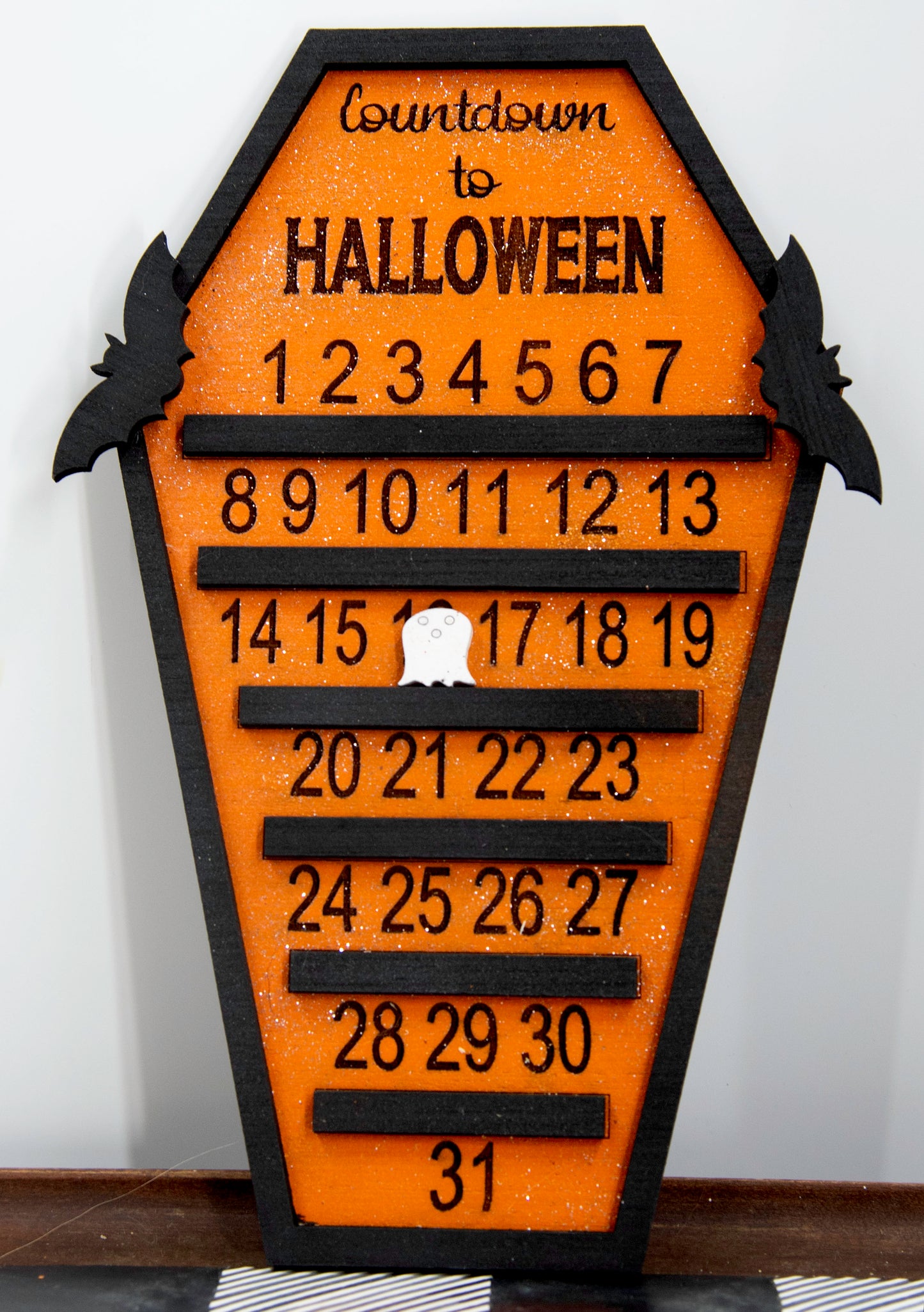 Wooden Halloween countdown sign shaped like a coffin with an orange glitter background, black trim frame, engraved 'Countdown to Halloween' at the top, engraved numbers 1–31 with small black shelves below each, and three interchangeable markers: ghost, witch hat, and pumpkin. Includes bat accents on top.