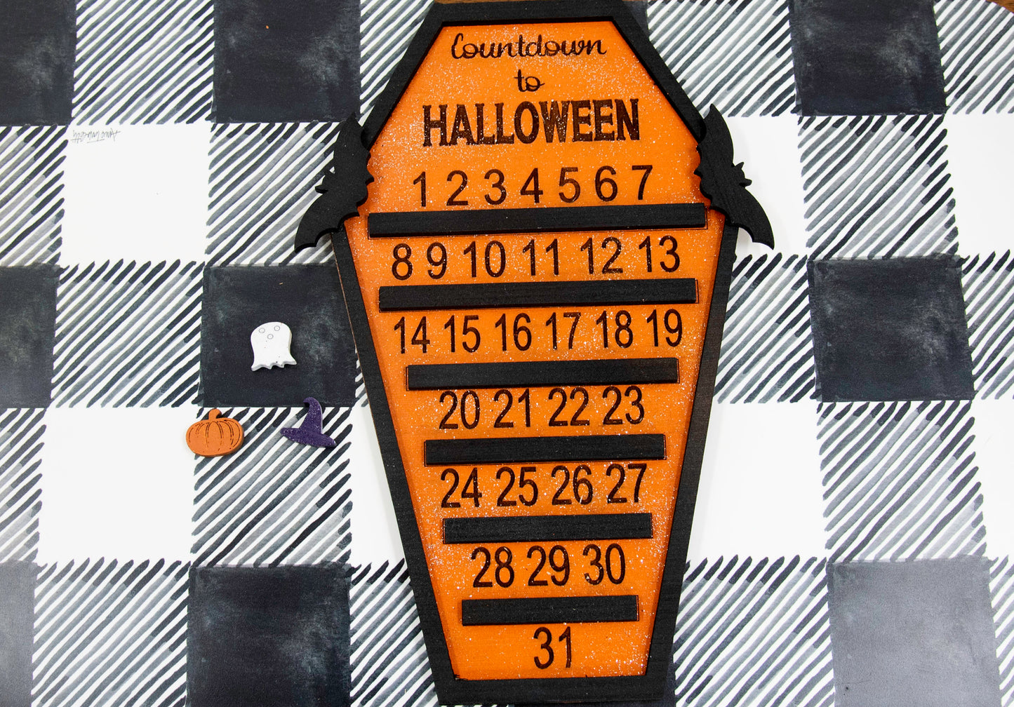Wooden Halloween countdown sign shaped like a coffin with an orange glitter background, black trim frame, engraved 'Countdown to Halloween' at the top, engraved numbers 1–31 with small black shelves below each, and three interchangeable markers: ghost, witch hat, and pumpkin. Includes bat accents on top.