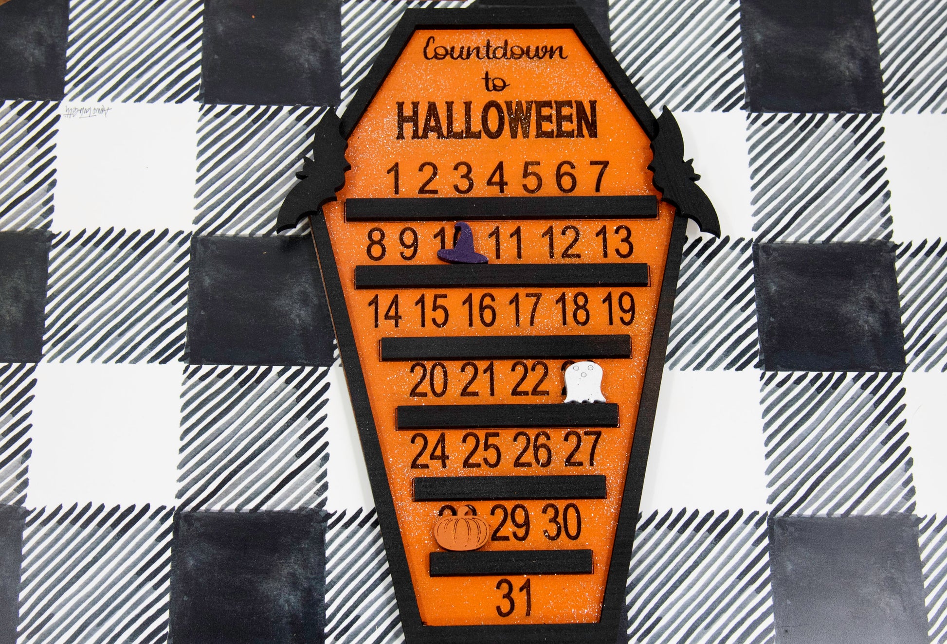 Wooden Halloween countdown sign shaped like a coffin with an orange glitter background, black trim frame, engraved 'Countdown to Halloween' at the top, engraved numbers 1–31 with small black shelves below each, and three interchangeable markers: ghost, witch hat, and pumpkin. Includes bat accents on top.