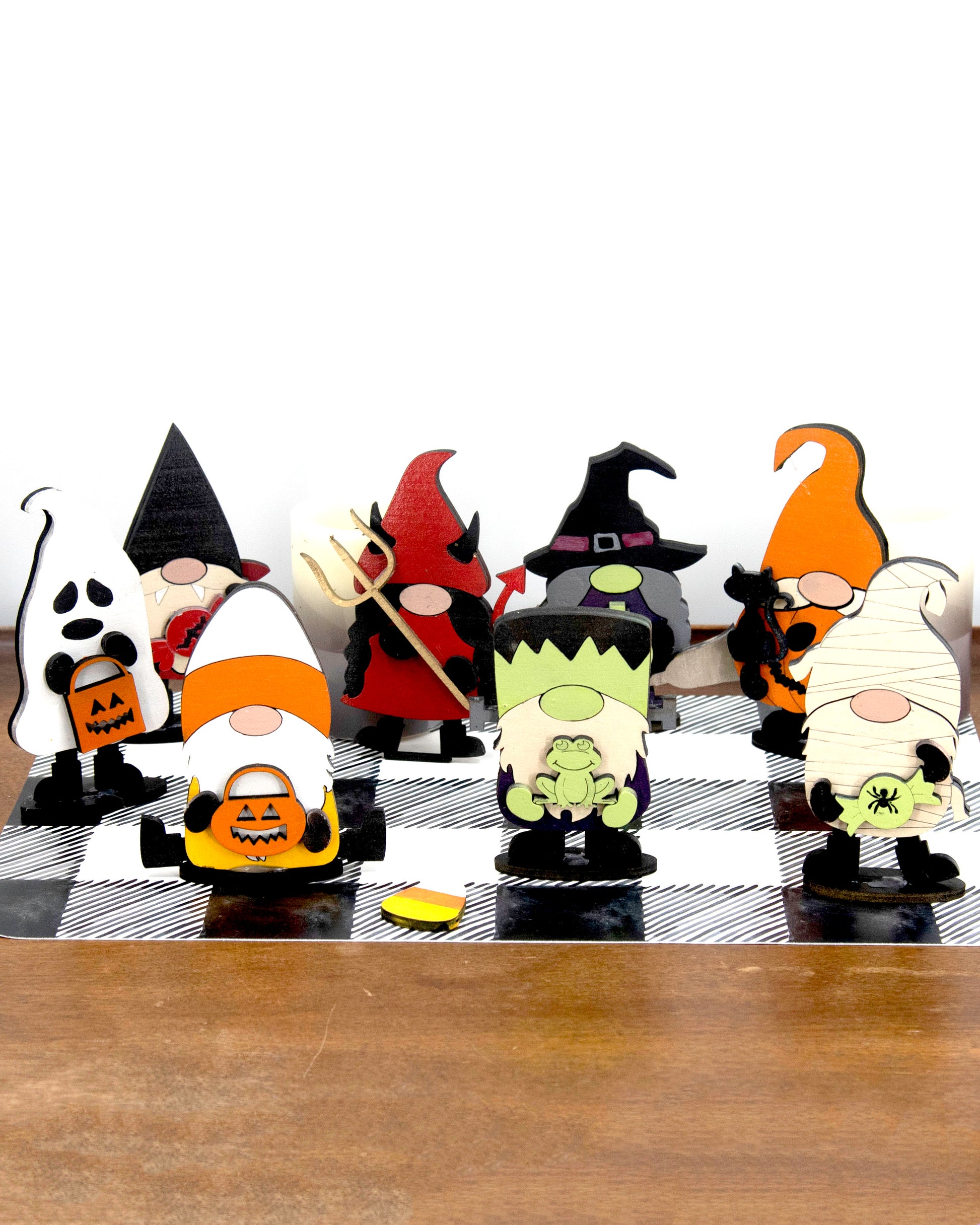 Set of hand-painted wooden Halloween gnome figurines dressed in spooky costumes including witch, ghost, pumpkin, vampire, and Frankenstein, standing on a shelf with black and white buffalo check runner.
