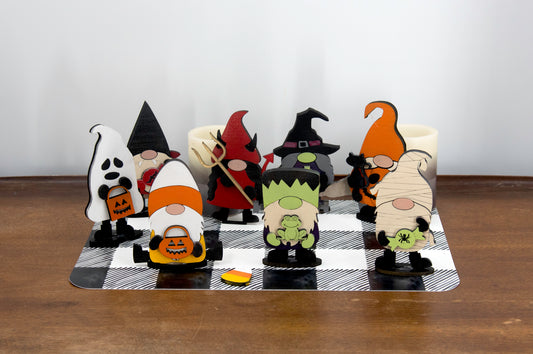 Set of hand-painted wooden Halloween gnome figurines dressed in spooky costumes including witch, ghost, pumpkin, vampire, and Frankenstein, standing on a shelf with black and white buffalo check runner.