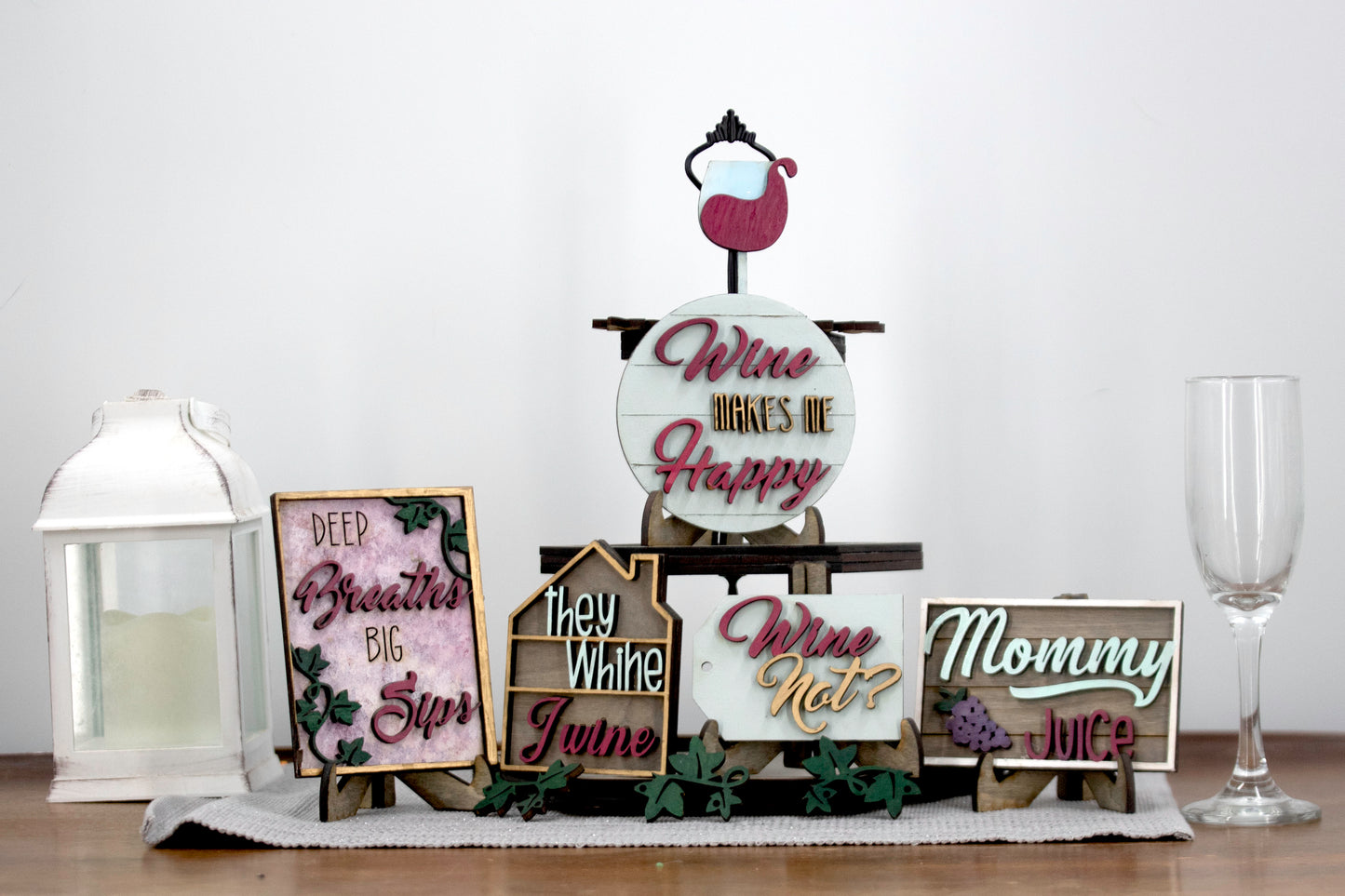A 6-piece wine-themed wooden tiered tray set featuring humorous mom quotes like "They whine, I wine" and "Mommy juice," painted in maroon, grey, gold, and light grey with included stands and optional tiered tray
