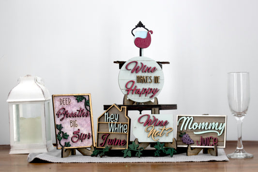 A 6-piece wine-themed wooden tiered tray set featuring humorous mom quotes like "They whine, I wine" and "Mommy juice," painted in maroon, grey, gold, and light grey with included stands and optional tiered tray
