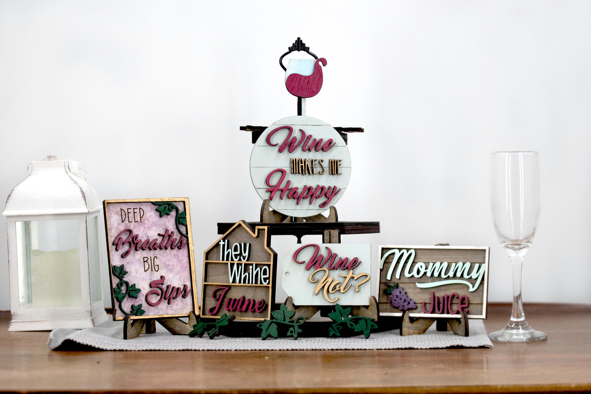A 6-piece wine-themed wooden tiered tray set featuring humorous mom quotes like "They whine, I wine" and "Mommy juice," painted in maroon, grey, gold, and light grey with included stands and optional tiered tray
