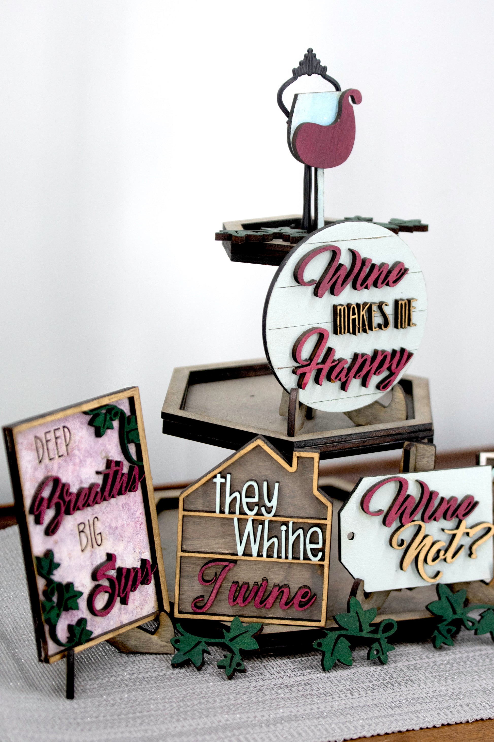 A 6-piece wine-themed wooden tiered tray set featuring humorous mom quotes like "They whine, I wine" and "Mommy juice," painted in maroon, grey, gold, and light grey with included stands and optional tiered tray
