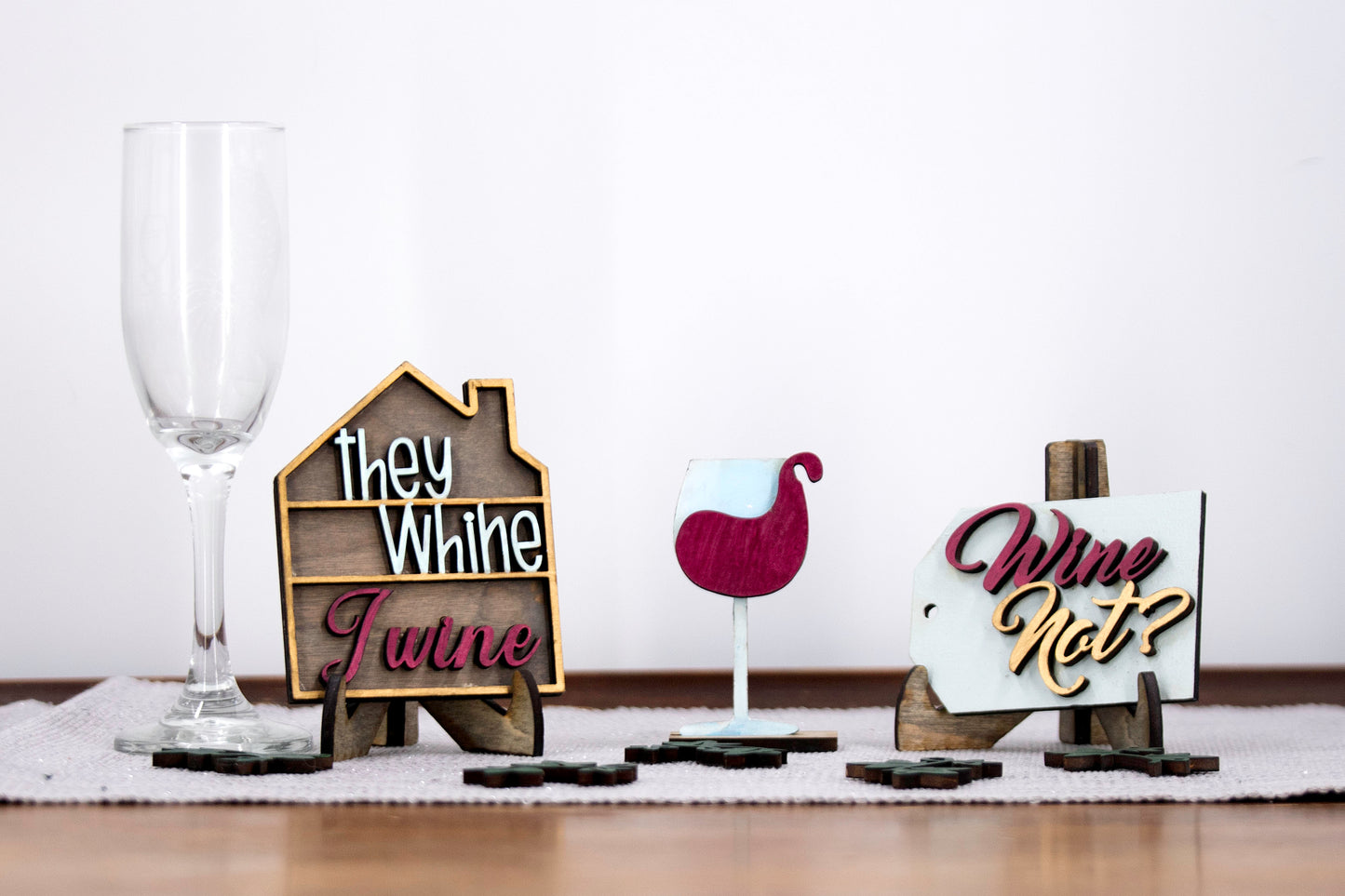 A 6-piece wine-themed wooden tiered tray set featuring humorous mom quotes like "They whine, I wine" and "Mommy juice," painted in maroon, grey, gold, and light grey with included stands and optional tiered tray
