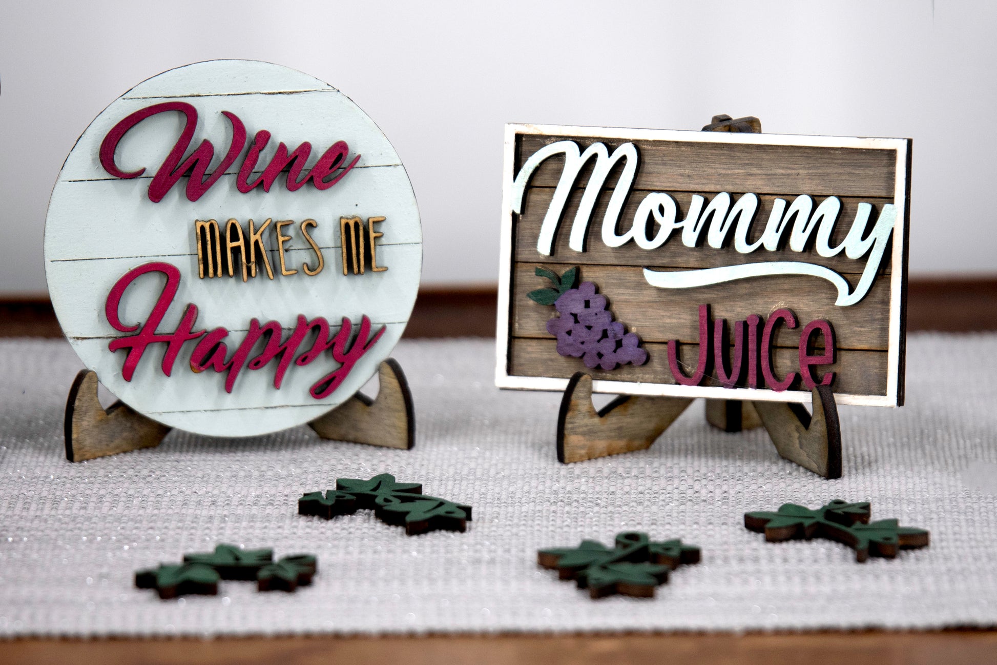 A 6-piece wine-themed wooden tiered tray set featuring humorous mom quotes like "They whine, I wine" and "Mommy juice," painted in maroon, grey, gold, and light grey with included stands and optional tiered tray
