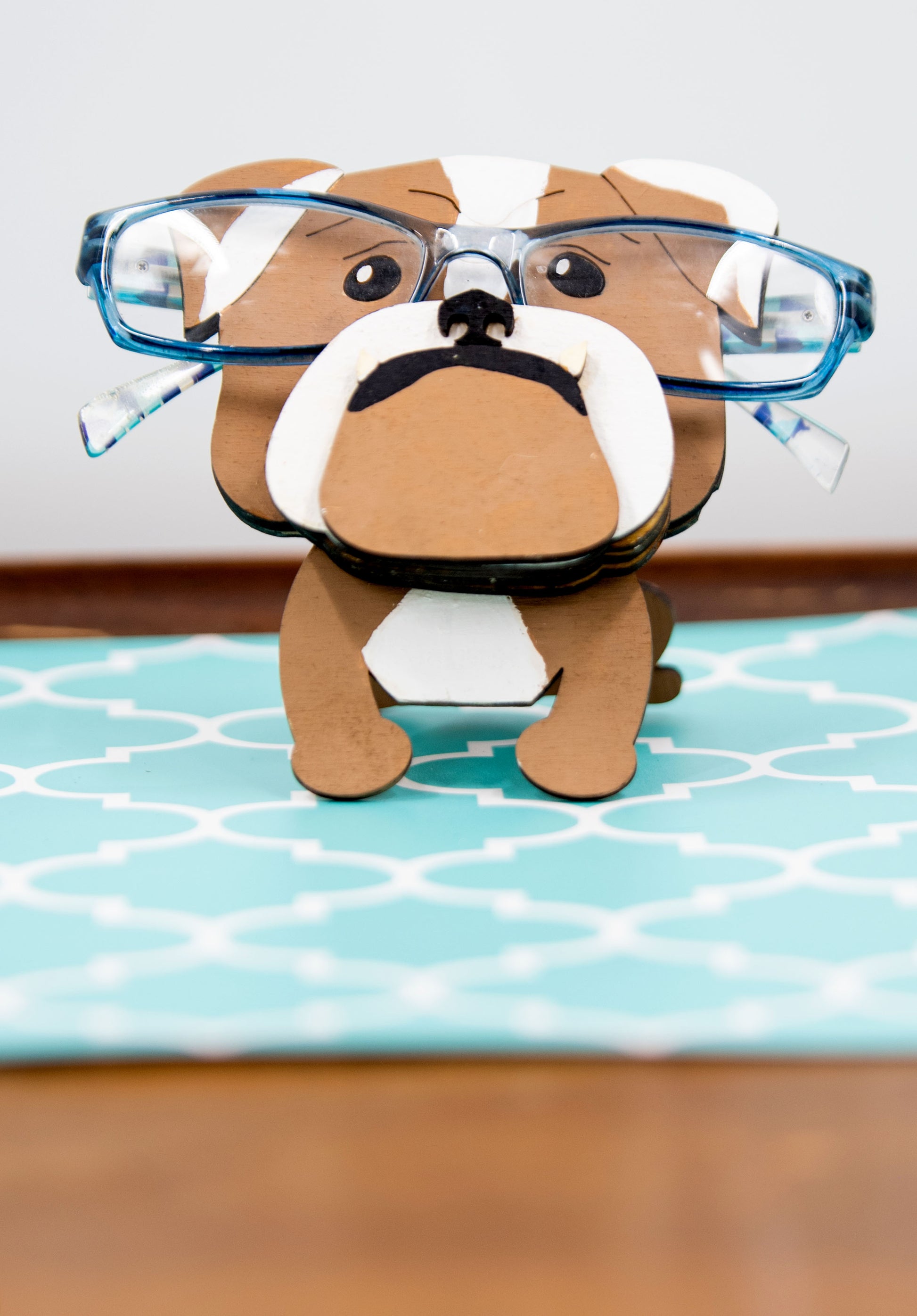 A hand-painted wooden glasses stand shaped like the face of an English Bulldog, featuring the breed’s signature wrinkled face and droopy cheeks. Designed with a flat base for stability, the stand holds eyeglasses securely on the Bulldog’s nose, making it a functional and charming decor piece for desks, nightstands, or shelves.