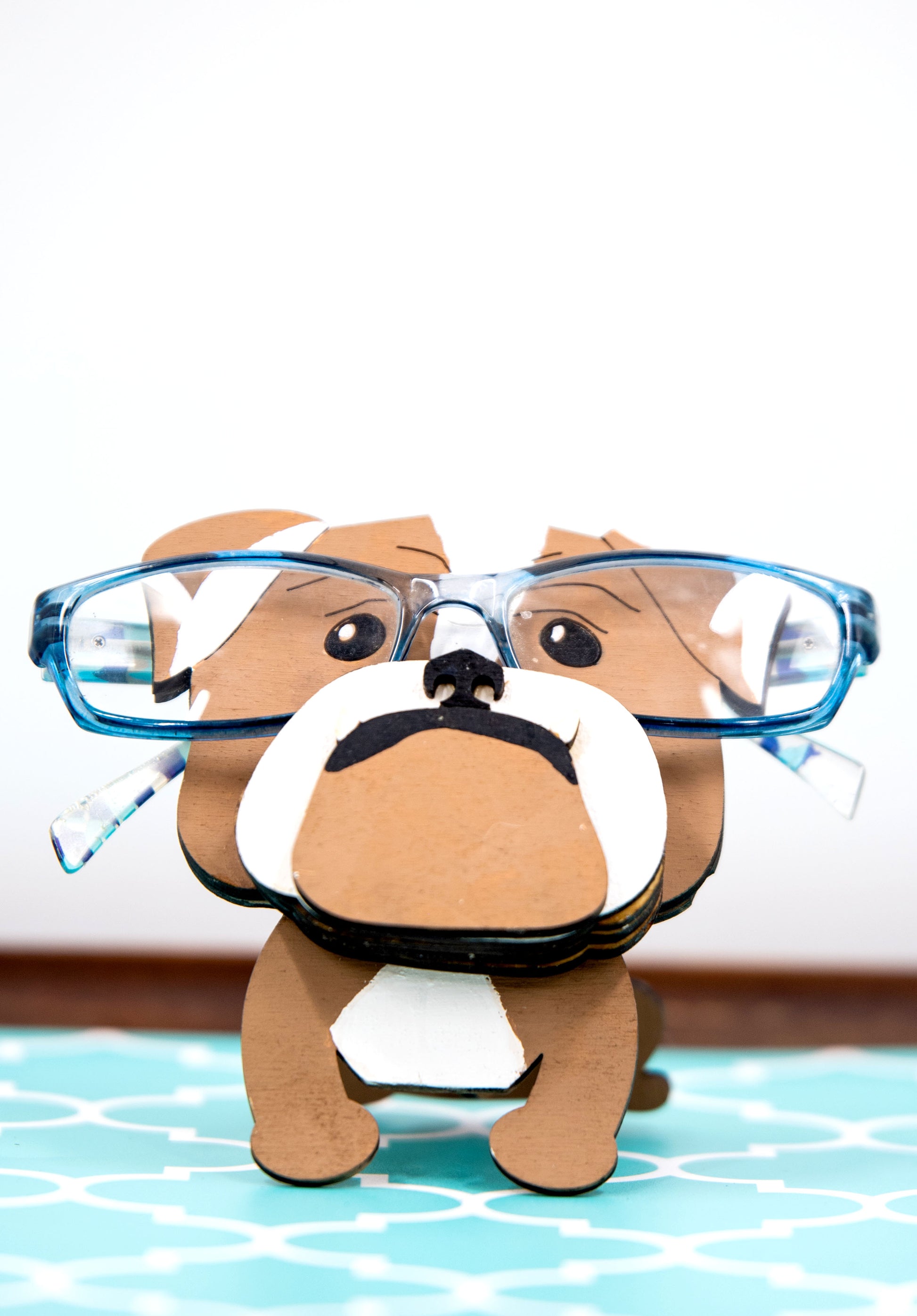 A hand-painted wooden glasses stand shaped like the face of an English Bulldog, featuring the breed’s signature wrinkled face and droopy cheeks. Designed with a flat base for stability, the stand holds eyeglasses securely on the Bulldog’s nose, making it a functional and charming decor piece for desks, nightstands, or shelves.