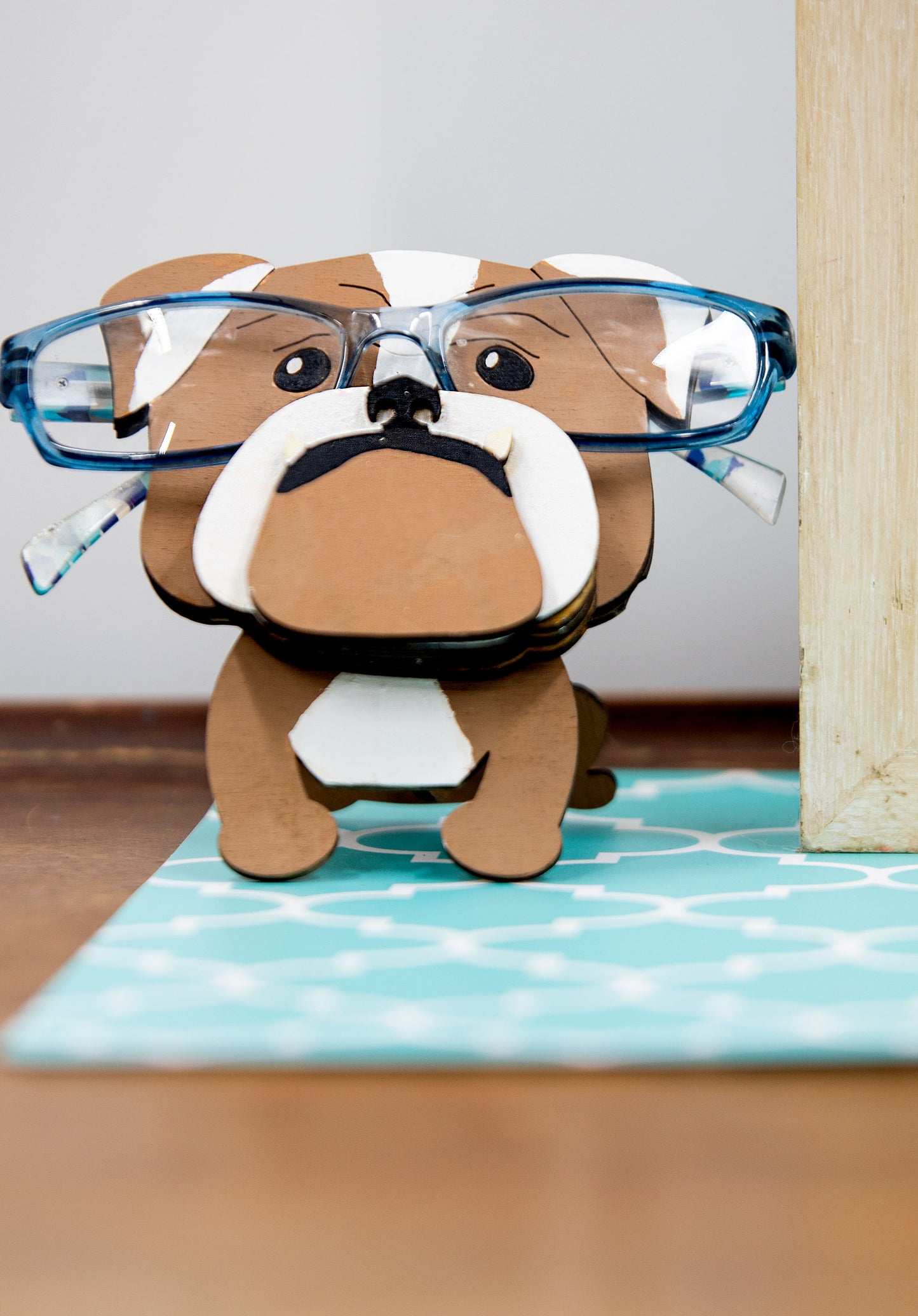 A hand-painted wooden glasses stand shaped like the face of an English Bulldog, featuring the breed’s signature wrinkled face and droopy cheeks. Designed with a flat base for stability, the stand holds eyeglasses securely on the Bulldog’s nose, making it a functional and charming decor piece for desks, nightstands, or shelves.
