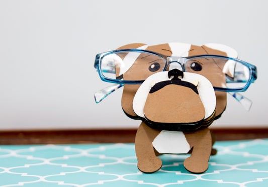 A hand-painted wooden glasses stand shaped like the face of an English Bulldog, featuring the breed’s signature wrinkled face and droopy cheeks. Designed with a flat base for stability, the stand holds eyeglasses securely on the Bulldog’s nose, making it a functional and charming decor piece for desks, nightstands, or shelves.
