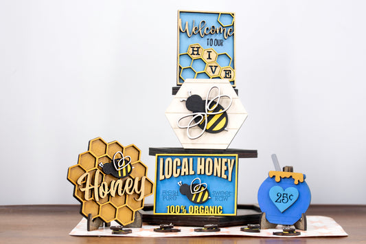Hand-painted bee tiered tray set with 6 summer-themed pieces including a honey jar, local honey sign, welcome to our hive sign, honey bomb, hexagon bee plaque, and 5 mini bees. Farmhouse style shelf sitters for kitchen, summer, or rustic decor. Finished and ready to display with optional matching tiered tray. Features custom honey bee designs, bright yellow accents, rustic wooden details, and custom color options. Perfect for seasonal tiered trays or bee-themed decor lovers.