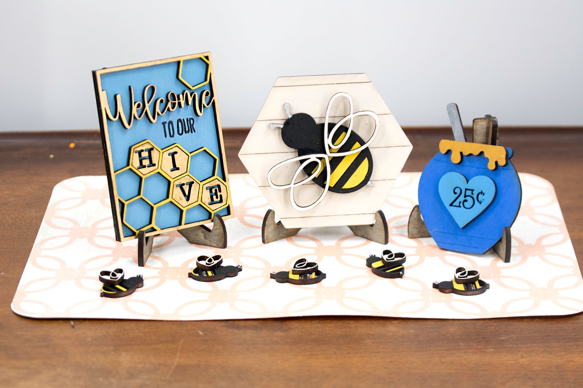 DIY bee tiered tray kit with 6 unfinished wood pieces including honey jar, local honey sign, welcome to our hive sign, honey bomb, bee hexagon plaque, and 5 small bee accents. Ideal for painting, staining, and customizing. Comes with stands for easy display. Perfect for summer crafts, farmhouse style decor, or paint-your-own projects. Great for hobby crafters, DIY parties, and seasonal decorating fun with a bee and honey theme.
