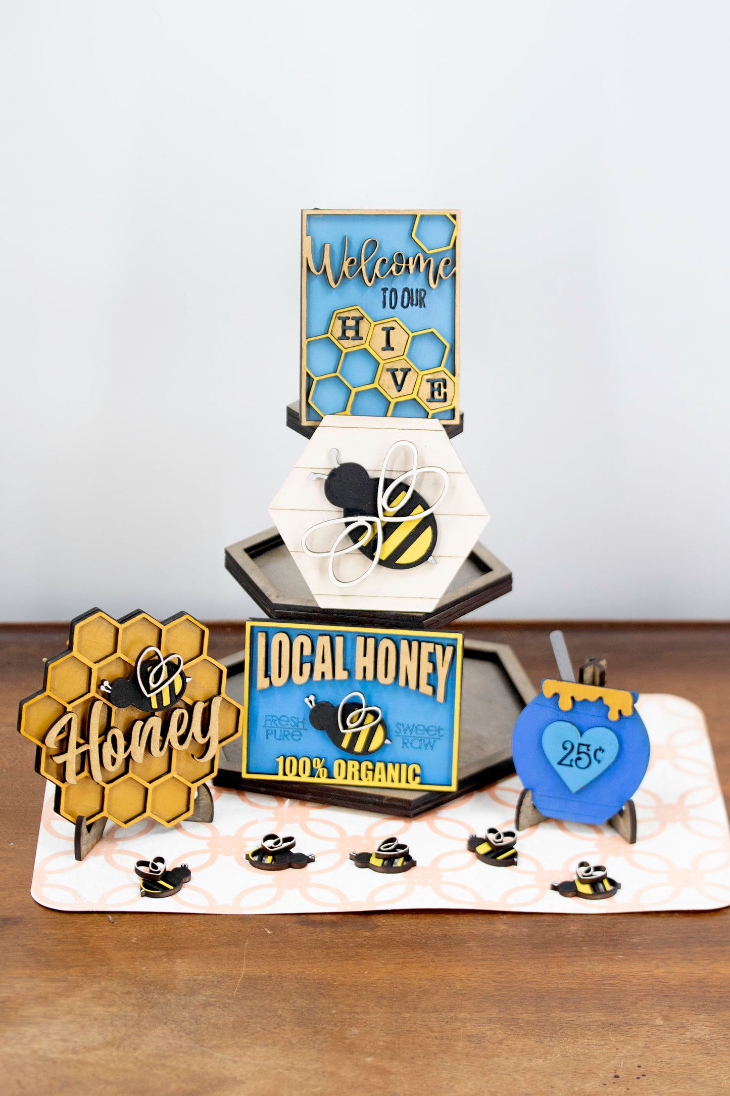 DIY bee tiered tray kit with 6 unfinished wood pieces including honey jar, local honey sign, welcome to our hive sign, honey bomb, bee hexagon plaque, and 5 small bee accents. Ideal for painting, staining, and customizing. Comes with stands for easy display. Perfect for summer crafts, farmhouse style decor, or paint-your-own projects. Great for hobby crafters, DIY parties, and seasonal decorating fun with a bee and honey theme.
