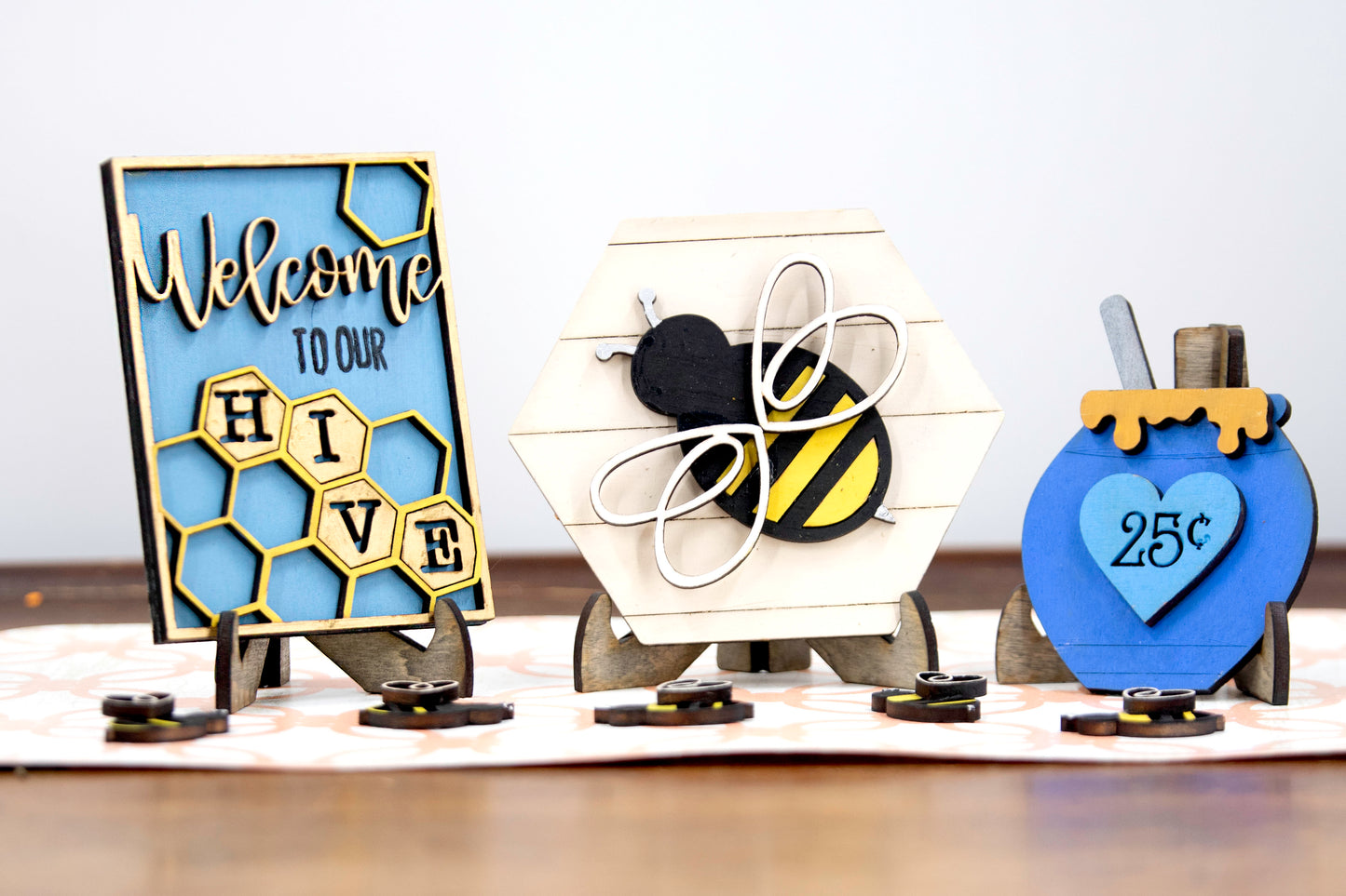 Hand-painted bee tiered tray set with 6 summer-themed pieces including a honey jar, local honey sign, welcome to our hive sign, honey bomb, hexagon bee plaque, and 5 mini bees. Farmhouse style shelf sitters for kitchen, summer, or rustic decor. Finished and ready to display with optional matching tiered tray. Features custom honey bee designs, bright yellow accents, rustic wooden details, and custom color options. Perfect for seasonal tiered trays or bee-themed decor lovers.