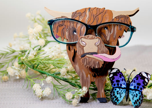 Wooden Highland Cow glasses holder with layered fur detail and curled horns, wearing eyeglasses and sticking out its tongue, surrounded by flowers and a decorative butterfly for a rustic and playful look.
