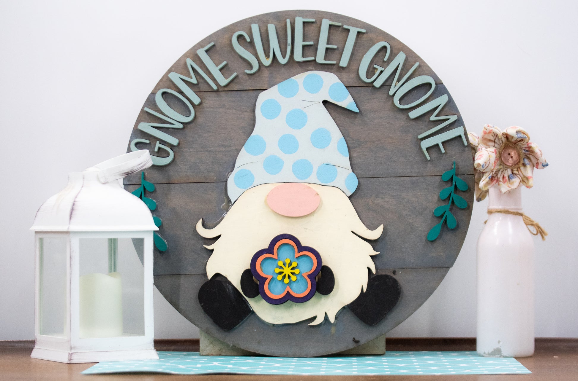 DIY kit for interchangeable gnome sweet gnome round sign with unfinished grey slatted wood base, engraved text, whimsical gnome figure, and 12 blank seasonal pieces with velcro for painting and assembly, farmhouse rustic decor craft project for entryway or door.
