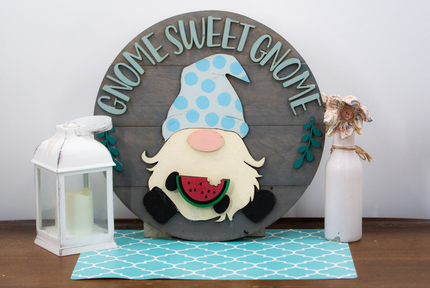 DIY kit for interchangeable gnome sweet gnome round sign with unfinished grey slatted wood base, engraved text, whimsical gnome figure, and 12 blank seasonal pieces with velcro for painting and assembly, farmhouse rustic decor craft project for entryway or door.
