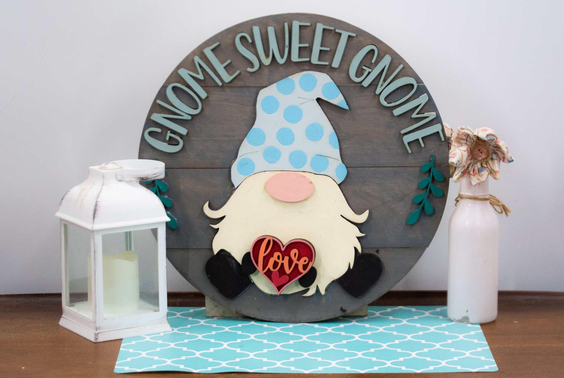 DIY kit for interchangeable gnome sweet gnome round sign with unfinished grey slatted wood base, engraved text, whimsical gnome figure, and 12 blank seasonal pieces with velcro for painting and assembly, farmhouse rustic decor craft project for entryway or door.
