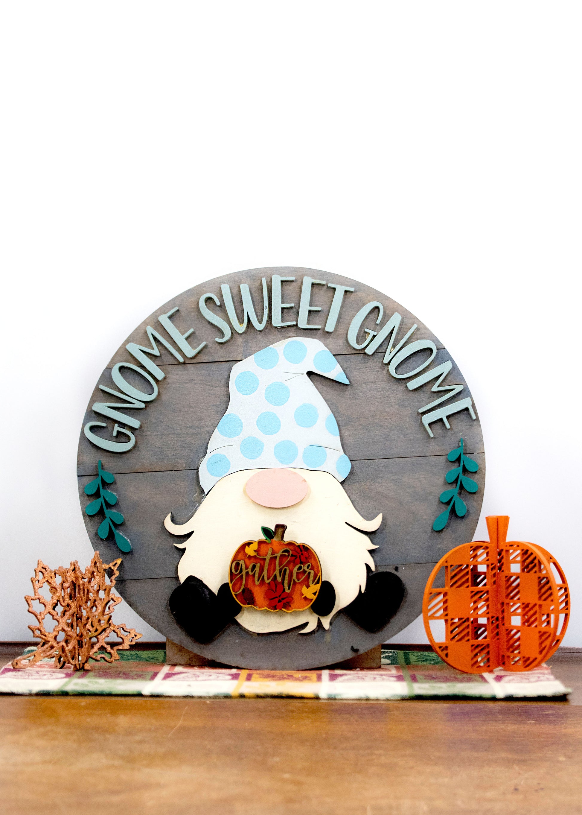 DIY kit for interchangeable gnome sweet gnome round sign with unfinished grey slatted wood base, engraved text, whimsical gnome figure, and 12 blank seasonal pieces with velcro for painting and assembly, farmhouse rustic decor craft project for entryway or door.
