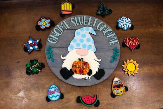 DIY kit for interchangeable gnome sweet gnome round sign with unfinished grey slatted wood base, engraved text, whimsical gnome figure, and 12 blank seasonal pieces with velcro for painting and assembly, farmhouse rustic decor craft project for entryway or door.
