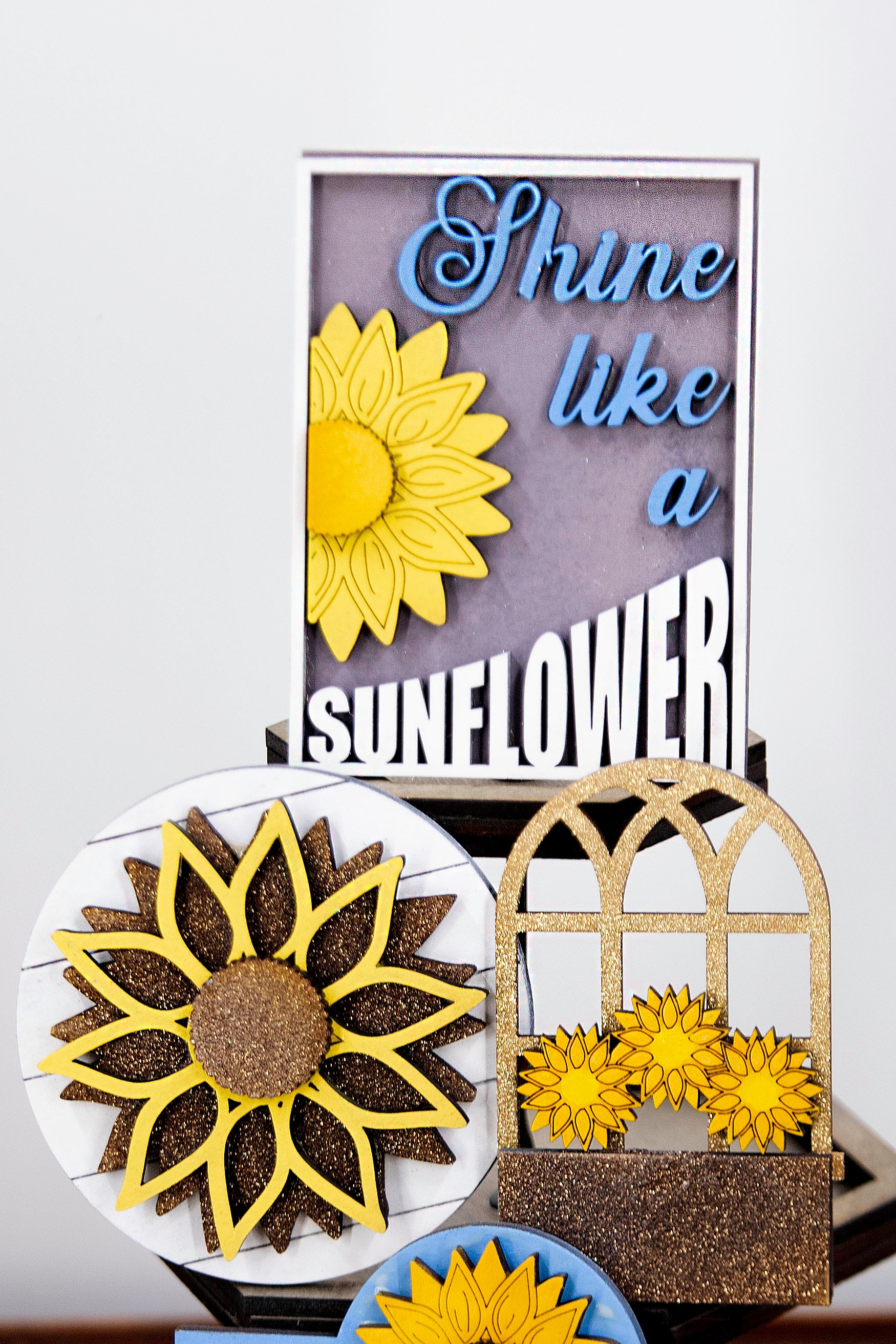 Sunflower-themed tiered tray set with six detailed pieces including signs like “Shine Like a Sunflower,” “Fresh Cut Sunflowers,” and “U Pick.” Includes a 6" faux slatted round with a sunflower graphic, window box, sweater weather sign, and five mini sunflower accents. Rustic farmhouse style in bright yellows and soft neutrals. Optional hexagon tray available. Perfect for fall or summer seasonal decor and kitchen styling.
