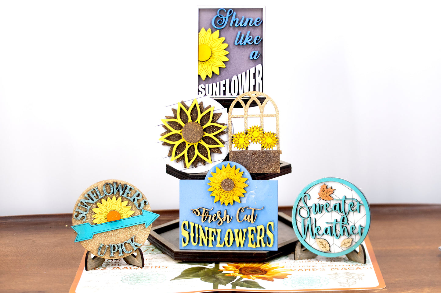 Sunflower-themed tiered tray set with six detailed pieces including signs like “Shine Like a Sunflower,” “Fresh Cut Sunflowers,” and “U Pick.” Includes a 6" faux slatted round with a sunflower graphic, window box, sweater weather sign, and five mini sunflower accents. Rustic farmhouse style in bright yellows and soft neutrals. Optional hexagon tray available. Perfect for fall or summer seasonal decor and kitchen styling.
