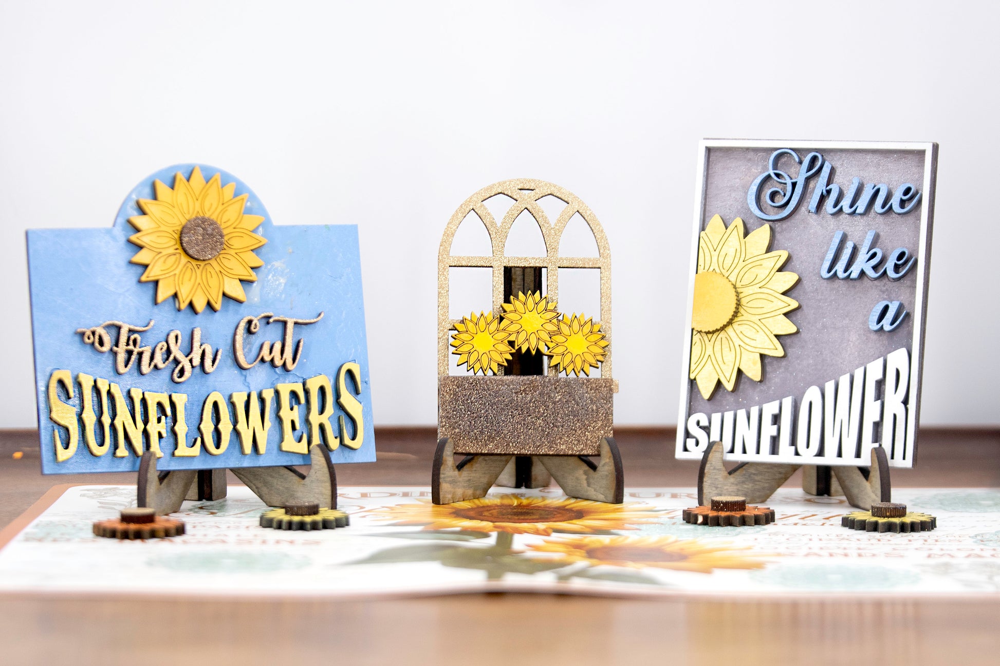 Sunflower-themed tiered tray set with six detailed pieces including signs like “Shine Like a Sunflower,” “Fresh Cut Sunflowers,” and “U Pick.” Includes a 6" faux slatted round with a sunflower graphic, window box, sweater weather sign, and five mini sunflower accents. Rustic farmhouse style in bright yellows and soft neutrals. Optional hexagon tray available. Perfect for fall or summer seasonal decor and kitchen styling.
