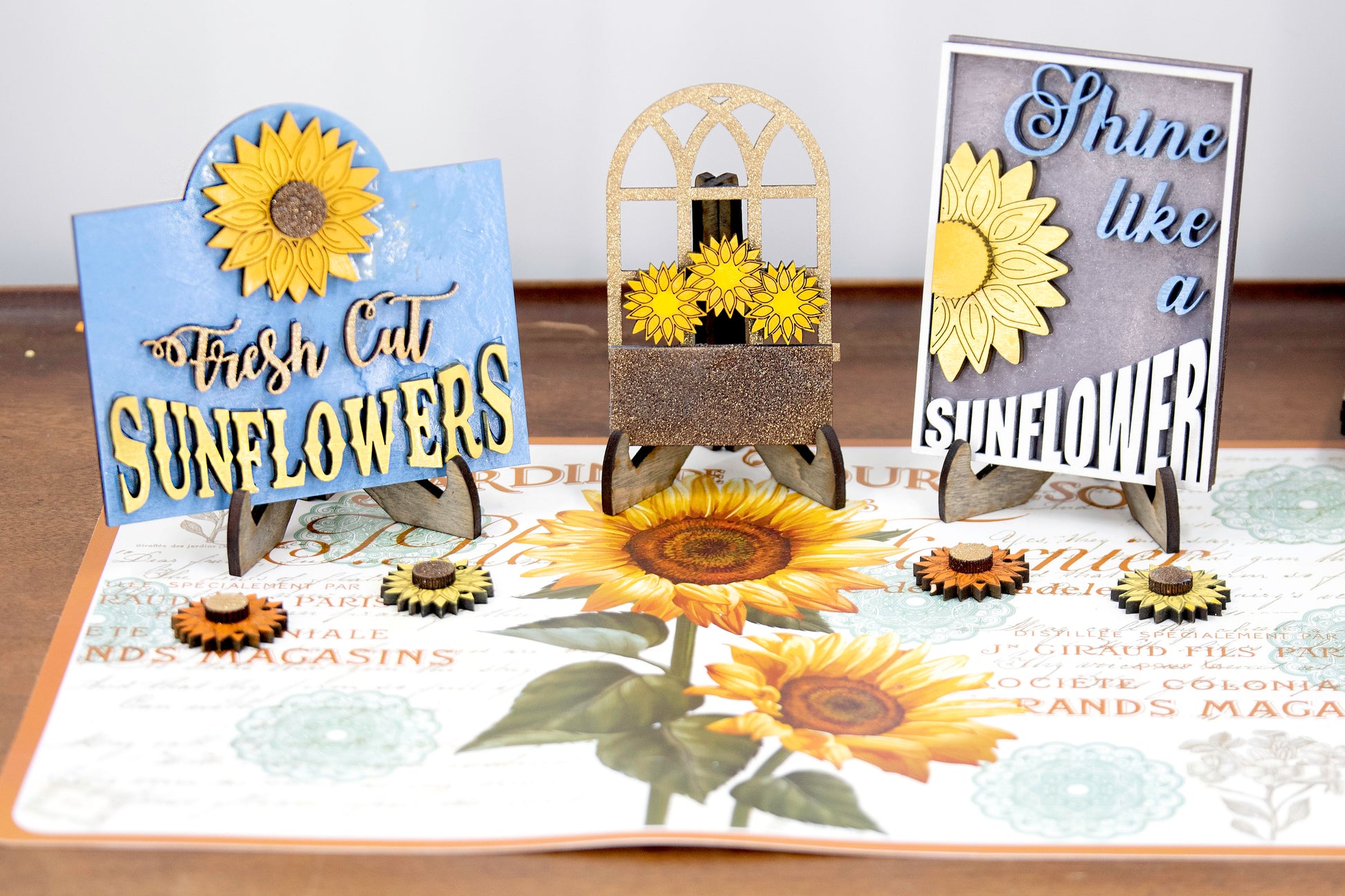 Sunflower-themed tiered tray set with six detailed pieces including signs like “Shine Like a Sunflower,” “Fresh Cut Sunflowers,” and “U Pick.” Includes a 6" faux slatted round with a sunflower graphic, window box, sweater weather sign, and five mini sunflower accents. Rustic farmhouse style in bright yellows and soft neutrals. Optional hexagon tray available. Perfect for fall or summer seasonal decor and kitchen styling.
