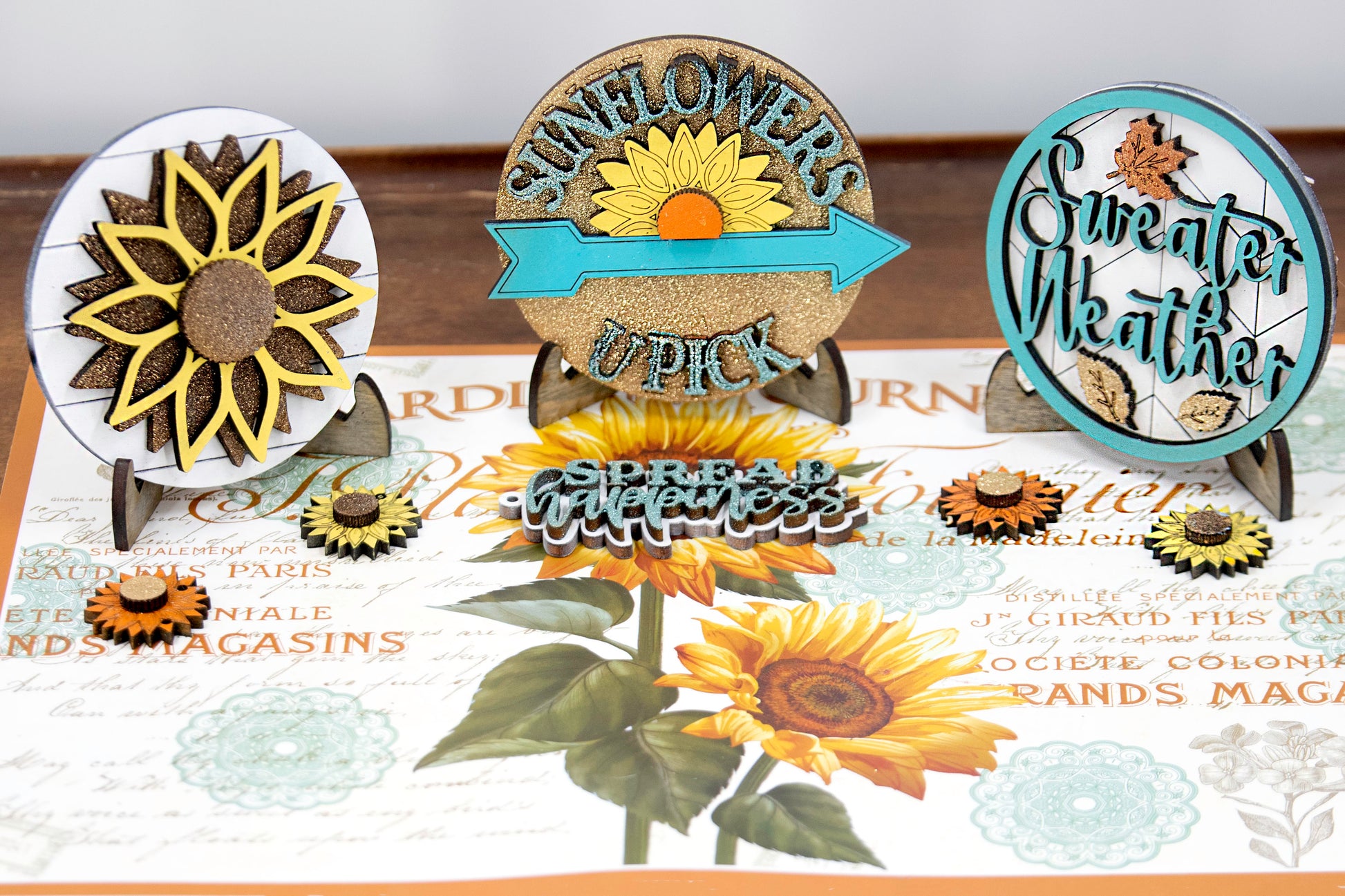 Sunflower-themed tiered tray set with six detailed pieces including signs like “Shine Like a Sunflower,” “Fresh Cut Sunflowers,” and “U Pick.” Includes a 6" faux slatted round with a sunflower graphic, window box, sweater weather sign, and five mini sunflower accents. Rustic farmhouse style in bright yellows and soft neutrals. Optional hexagon tray available. Perfect for fall or summer seasonal decor and kitchen styling.

