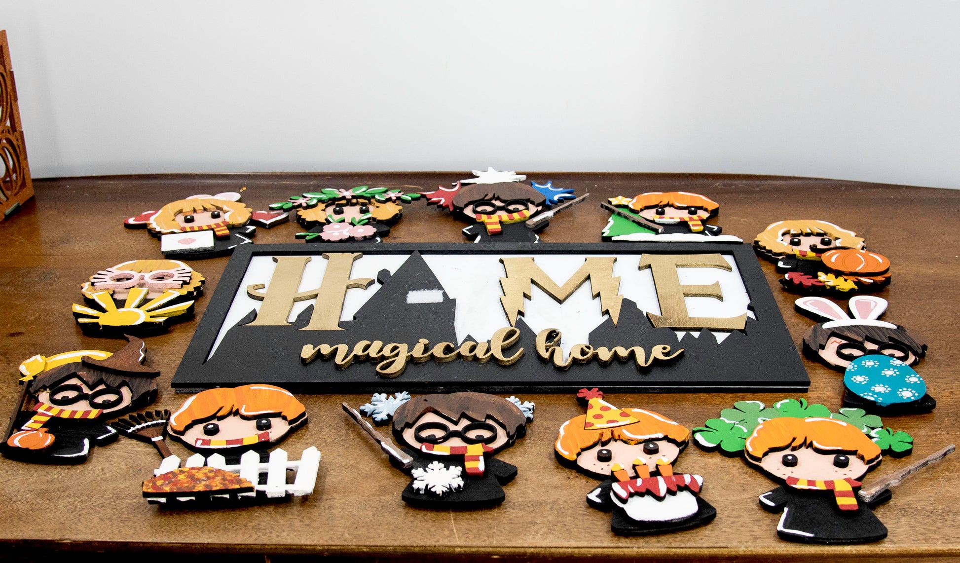 Full view of handcrafted wizard-themed interchangeable wooden sign that reads 'Home Magical Home' with wizard silhouettes forming the letter O, displayed on a shelf with all 12 hand-painted seasonal pieces including pumpkin, snowflake, shamrock, heart, sun, watermelon, leaf, ornament, egg, flower, flag, and candy corn — perfect for year-round magical home decor, Harry Potter-inspired, fantasy cottagecore aesthetic, unique seasonal gift for wizard fans and DIY crafters