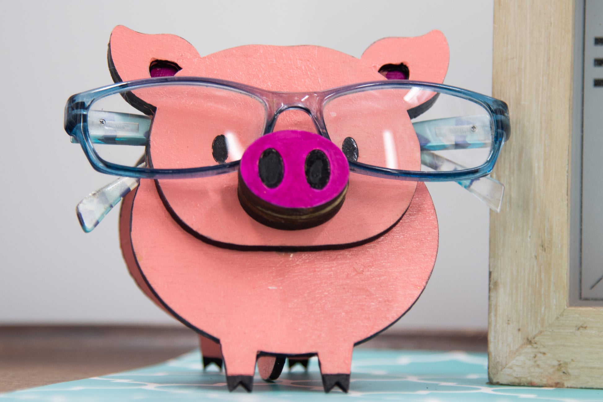Handcrafted wooden pig-shaped eyeglass stand with pink finish and black details, holding glasses on its snout, ideal for desks or gift giving.