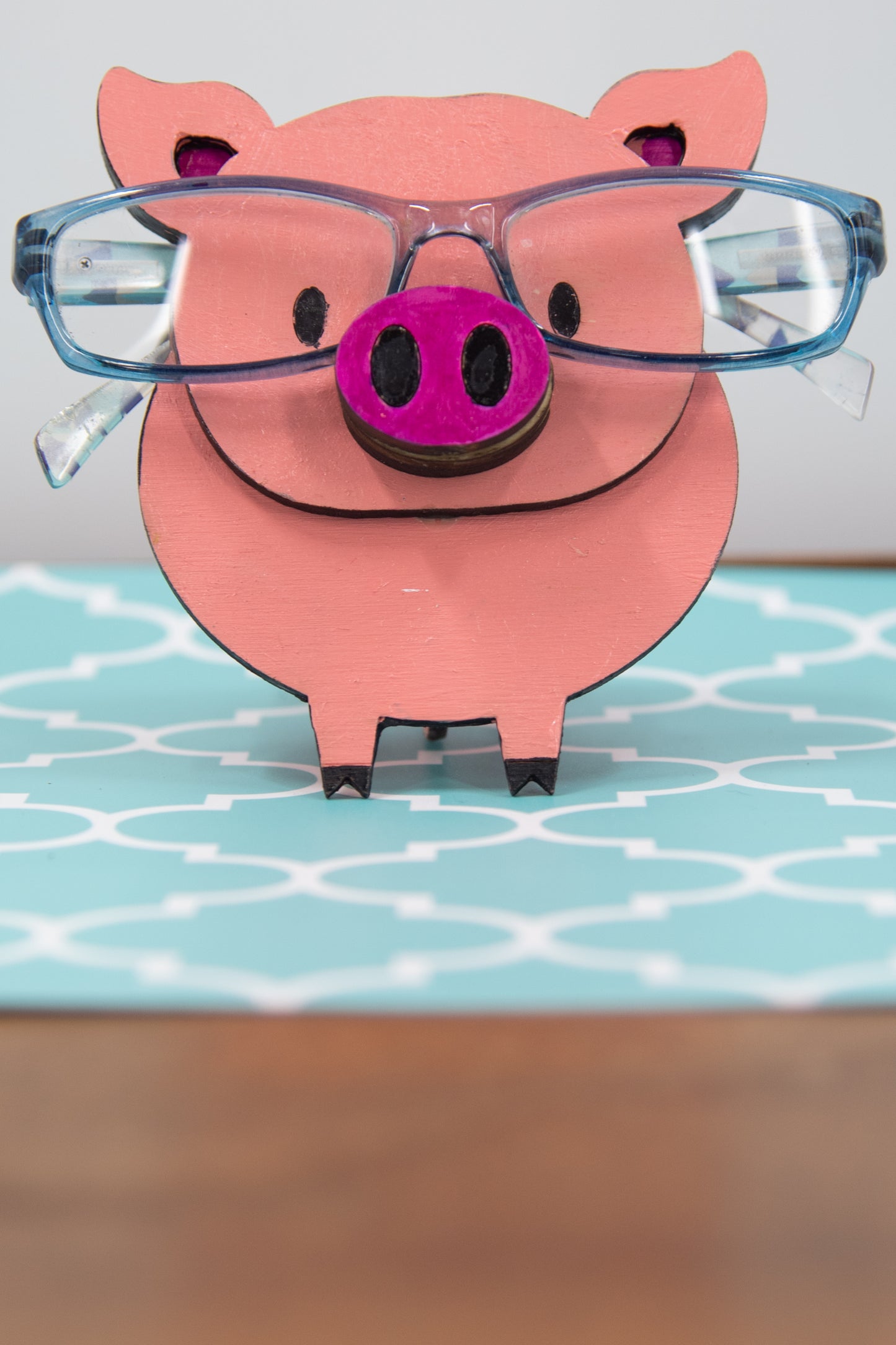 Handcrafted wooden pig-shaped eyeglass stand with pink finish and black details, holding glasses on its snout, ideal for desks or gift giving.